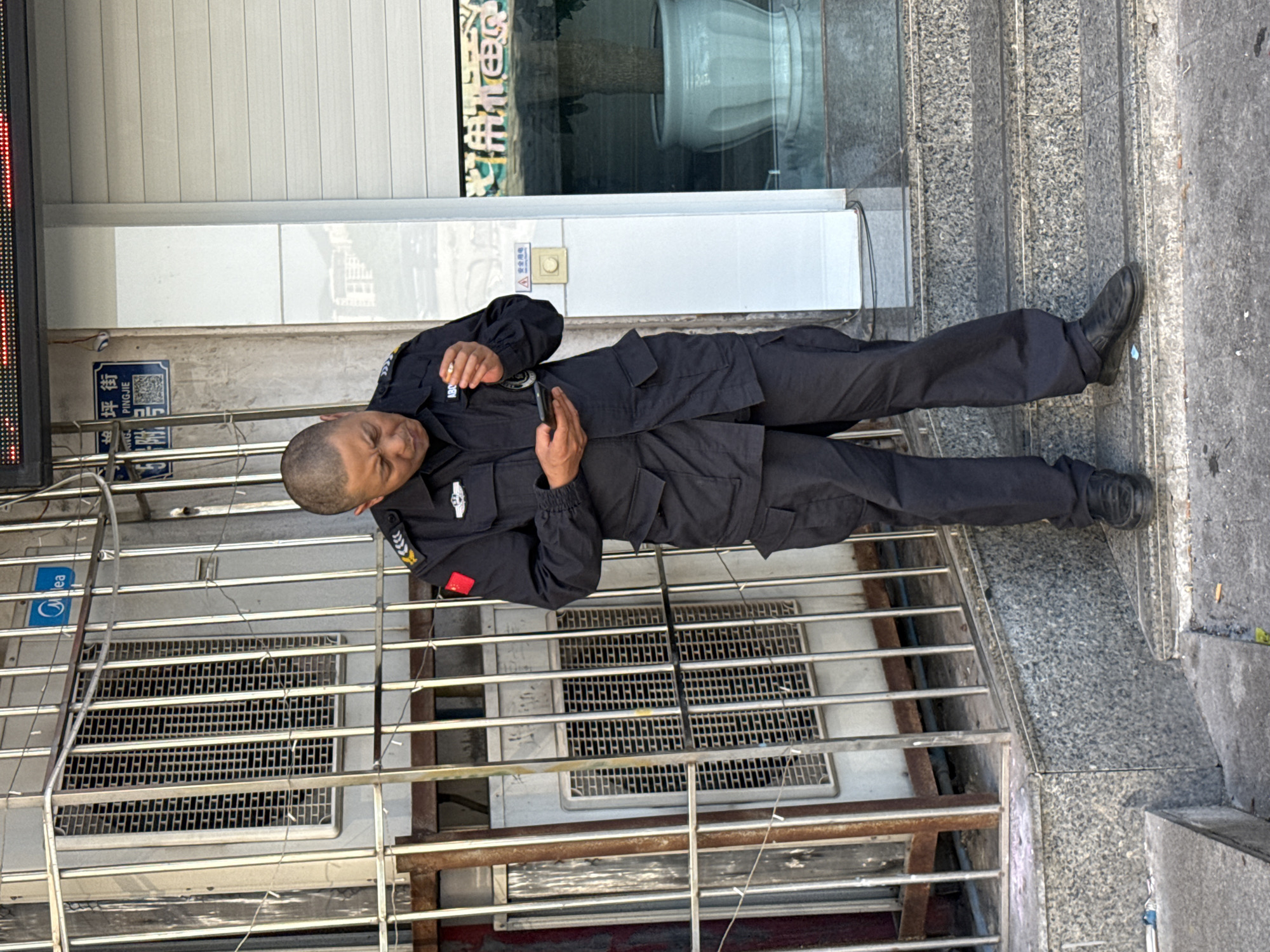 A man in a dark uniform stands near a metal grate outside a building with a gray marble step. He holds a cigarette and a mobile phone. A blue street sign, with Chinese characters is affixed to the exterior wall. The location is Jiulongpo District, Chongqing, China, based on the street sign that reads 'Pingjie.'