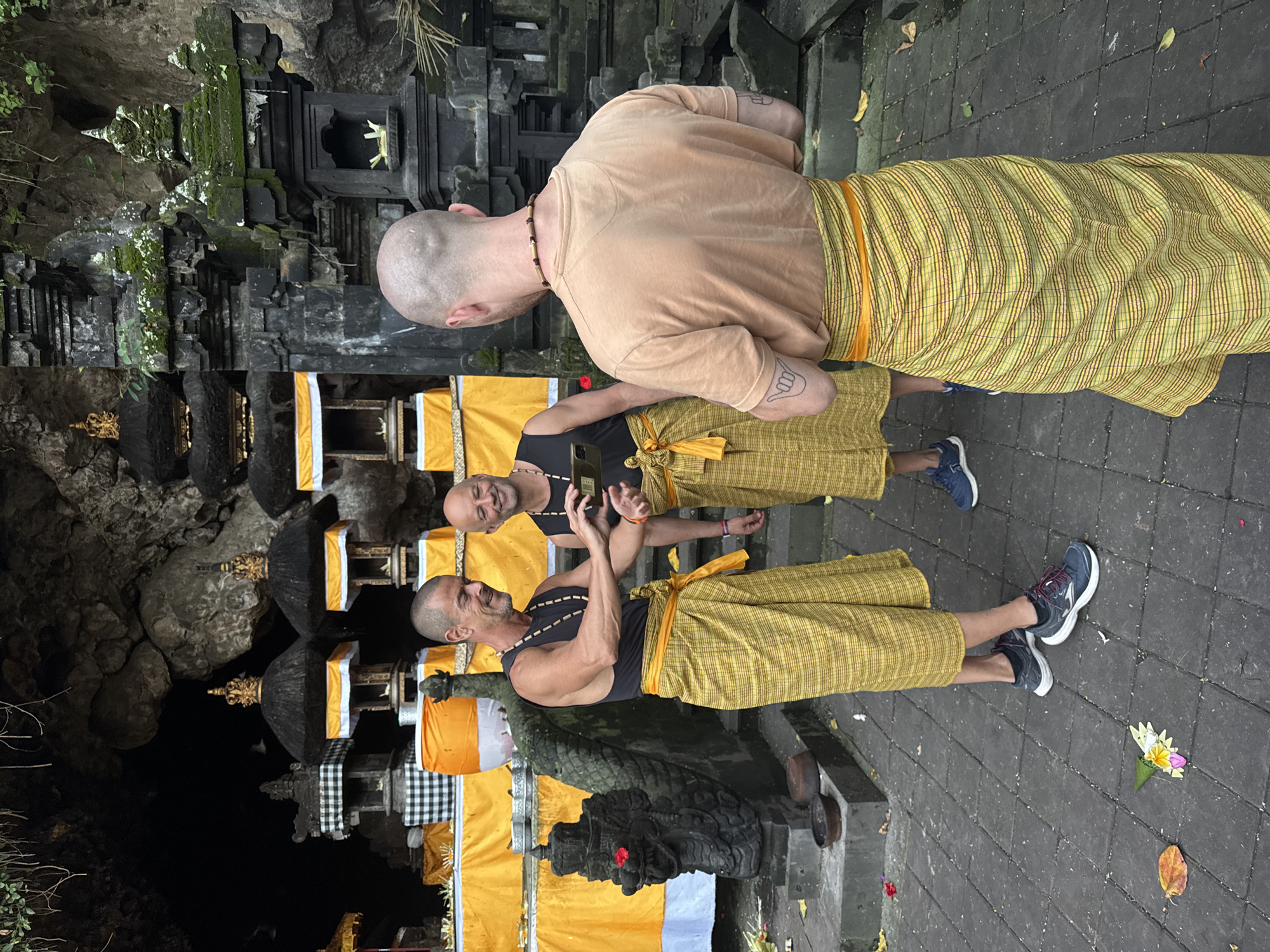 The image depicts three bald men in an outdoor setting with ornate stone structures and vibrant yellow fabric. The man on the left, wearing a black tank top and a yellow sarong, is holding a dark object, possibly a phone, and appears to be showing it to the man in the center. The man in the center, also in a black tank top and yellow sarong, is smiling and looking at the object. The man on the right, facing away from the camera, wears a light brown t-shirt and a yellow sash, and has a tattoo on his forearm. Behind them, there are several tiered structures adorned with dark roofs and golden spires, draped with yellow cloth and white and black checkered fabric. A stone serpent statue is visible on the left. The ground is paved with grey stone tiles. The background features a dark cave entrance and lush greenery overhead. This scene is located at Goa Gajah (Elephant Cave) in Dawan, Indonesia, a significant archaeological site and tourist attraction.