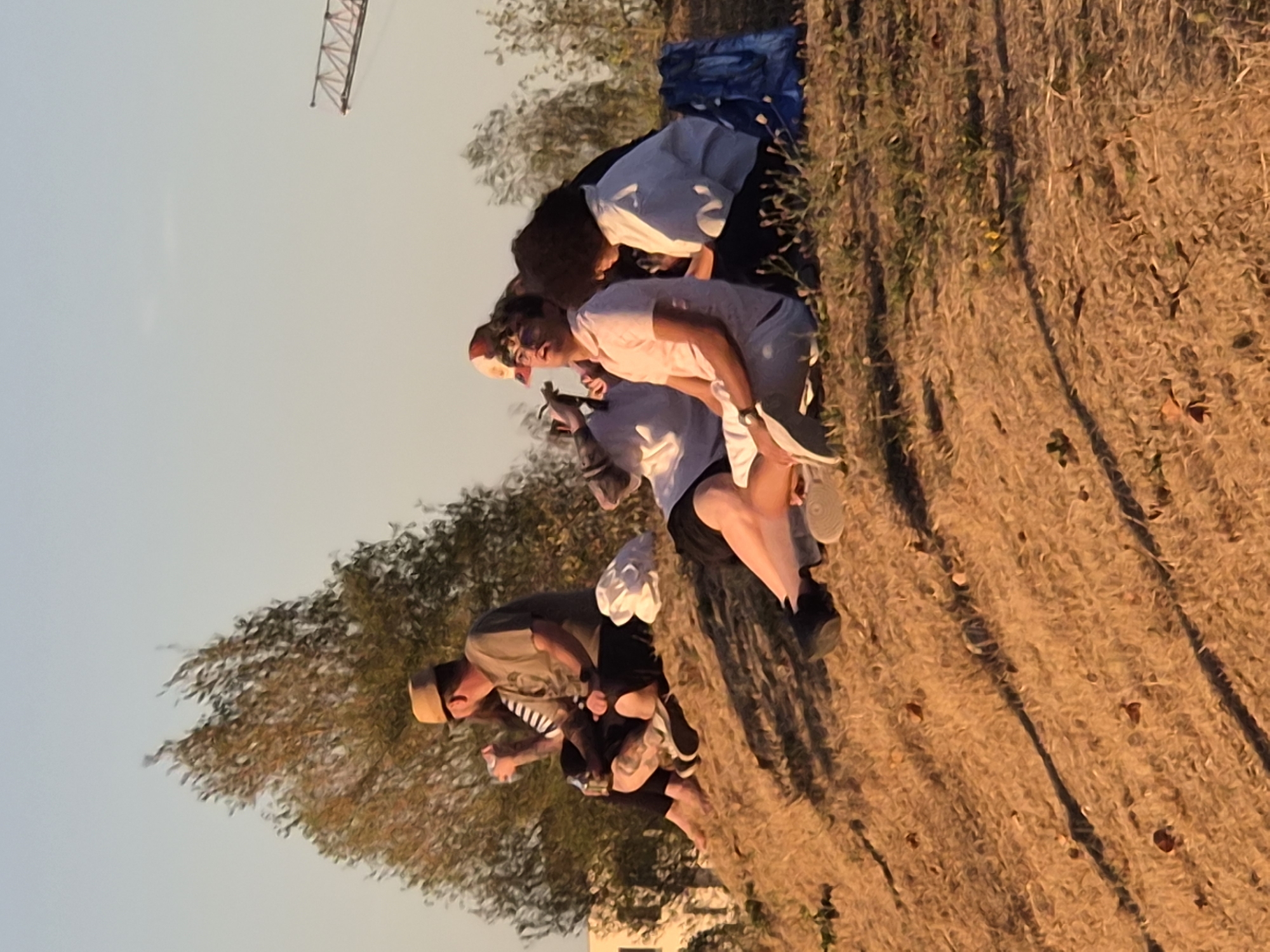 In Sanem, Luxembourg, a group of people are gathered on a grassy hill under a clear sky. The sun is low, casting long shadows and bathing the scene in golden light. A man in a tan hat and a green t-shirt sits with his arm around a woman in a striped top. To their right, a man in a pink shirt and white shorts is looking upwards, while a woman in a white shirt sits beside him, looking down. A blue bag rests on the ground near them. In the background, trees and a construction crane are visible. The overall atmosphere appears relaxed and casual.
