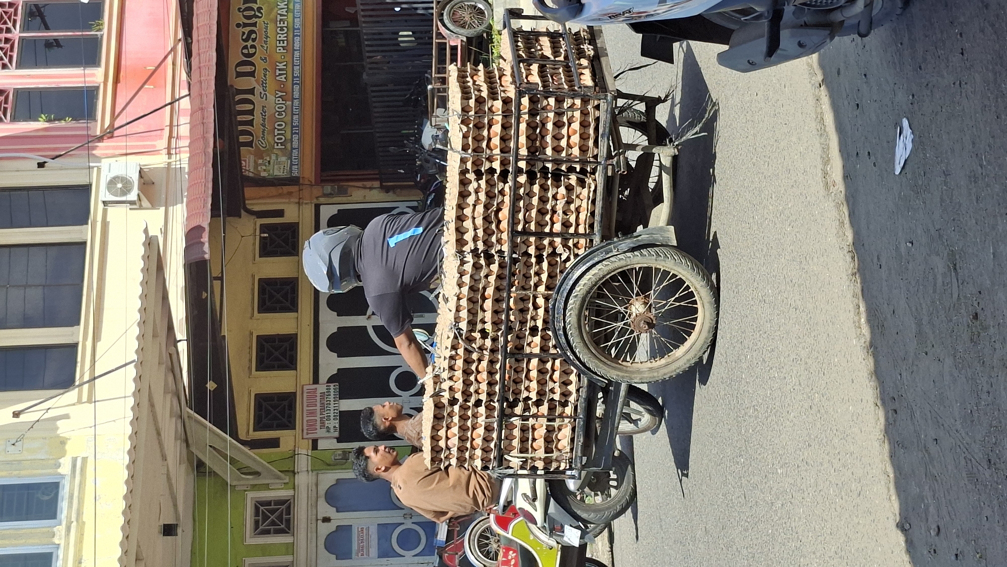 In Lhokseumawe, Indonesia, a man wearing a helmet and a black shirt with a blue number "1" on the back is operating a three-wheeled cart loaded with numerous stacks of egg cartons. He is traveling on a paved road in front of shops. Two other men are visible on a white motorcycle next to the cart. The shop in the background has signs that read "TOKO INI DIJUAL" and "UDU Design". The scene appears to be a street in a commercial area.