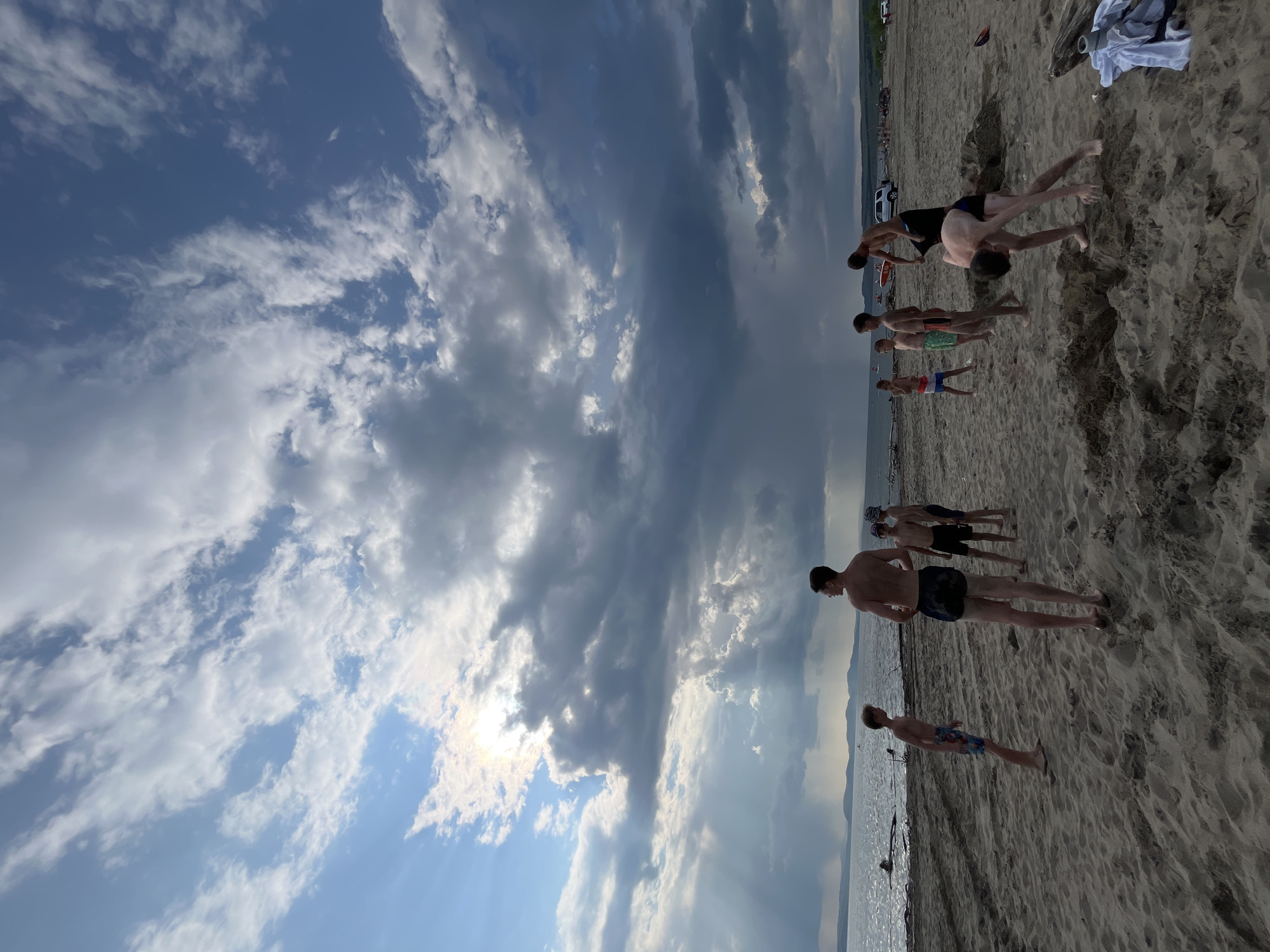 A group of young men are gathered on a sandy beach, possibly for an outdoor activity or sports. The scene is set under a dynamic sky with a mix of dark, dramatic clouds and brighter patches, suggesting the sun is either partially obscured or breaking through. The lighting indicates it could be late afternoon or early evening. The people are all wearing swim trunks, implying a warm weather setting and possibly a swim or water-related activity. Some individuals appear to be in motion, with one person in the foreground doing a handstand, and others in the background in what looks like a line, perhaps preparing for a race or drill. The beach is composed of fine sand, and the sea is visible in the background, reflecting the sky. Mountains or hills can be seen in the distance, providing a sense of the location's topography. The overall mood seems casual and active, typical of a beach gathering on a summer day. There is no visible text in the image.