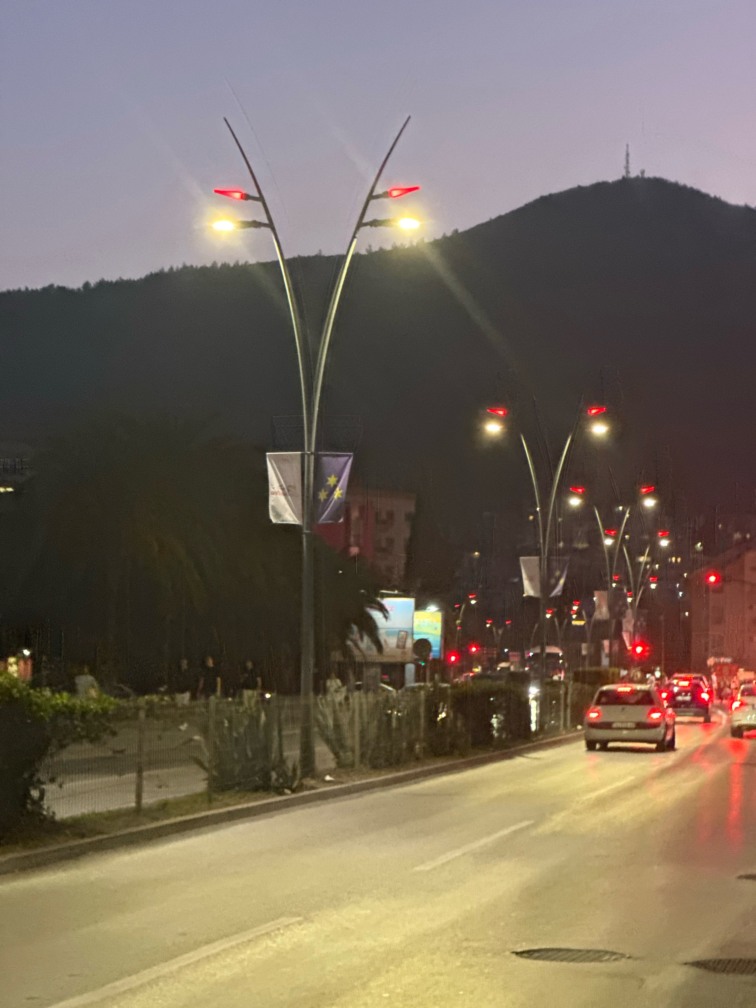 The image captures a street scene in Budva, Montenegro, at dusk. The sky is a muted purple and grey, indicating the transition from day to night. A dark, silhouetted hill with a telecommunications tower on its summit dominates the background. In the foreground, a street is illuminated by streetlights and the headlights and taillights of several cars. The streetlights have a distinct, modern design with curving metal arms holding multiple lights. Some of these lights are red, resembling traffic signals or decorative elements. Two banners hang from one of the lampposts; one is white with red text, and the other is purple with yellow stars, resembling the flag of the European Union.
Several cars are visible on the road, with their headlights and taillights glowing. One car, a light-colored sedan, is in the right lane, its brake lights illuminated. In the distance, more vehicles and traffic lights are visible, contributing to the urban atmosphere.
On the left side of the street, there is a pedestrian area with a fence and some greenery, including palm-like plants and bushes. A few people are standing or walking in this area, mostly silhouetted against the dim light. The overall impression is one of an active street at twilight, with the infrastructure of a European city illuminated as night falls.