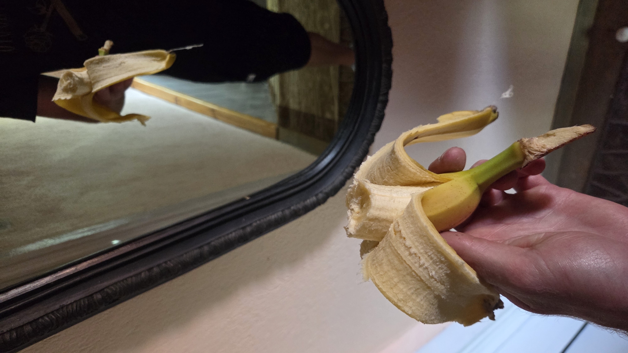 A person is holding a partially peeled banana. The banana is yellow with a green stem and appears to be fresh. The background shows a mirror reflecting another person holding a banana. The environment appears to be indoors, possibly a home or a public space. The lighting is dim.