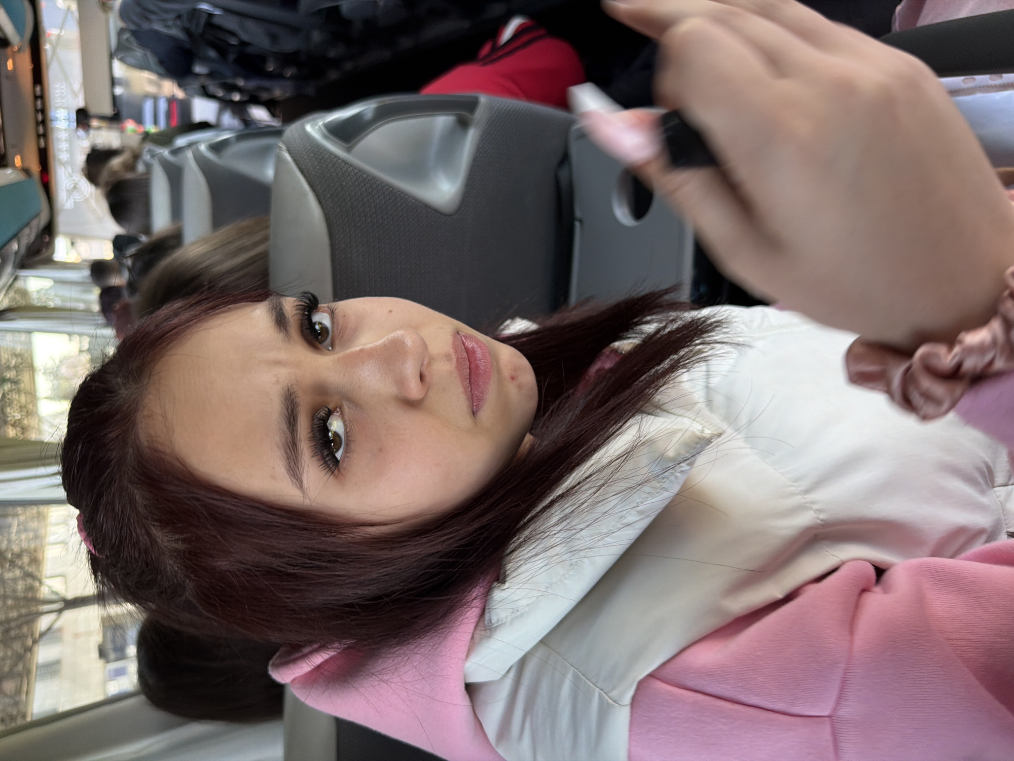 The image captures a young woman inside what appears to be a bus or train, looking directly towards the camera. Her head is tilted slightly. She has dark, likely auburn, hair, prominent long eyelashes, and light brown or hazel eyes, accentuated by light pink lipstick. She is wearing a white padded jacket over a pink hooded top. Her right hand is visible in the upper right corner of the frame, adorned with a pink scrunchie on her wrist, holding a dark, indistinct object.
In the background, rows of grey seats with headrests are visible, indicating a public transport vehicle. The backs of other passengers' heads can be seen further down the aisle. Through the windows on the left side, an outdoor scene is visible, featuring trees and buildings, suggesting daylight and an urban or suburban environment. Overhead racks with what looks like luggage or bags are also discernible.
The activity appears to be the woman posing for a photograph, possibly a selfie. The overall atmosphere suggests she is in transit. The time of day is daytime, evidenced by the light entering through the windows. No specific text is visible in the image. The location context for this scene is Kapitan Andreevo, Bulgaria.