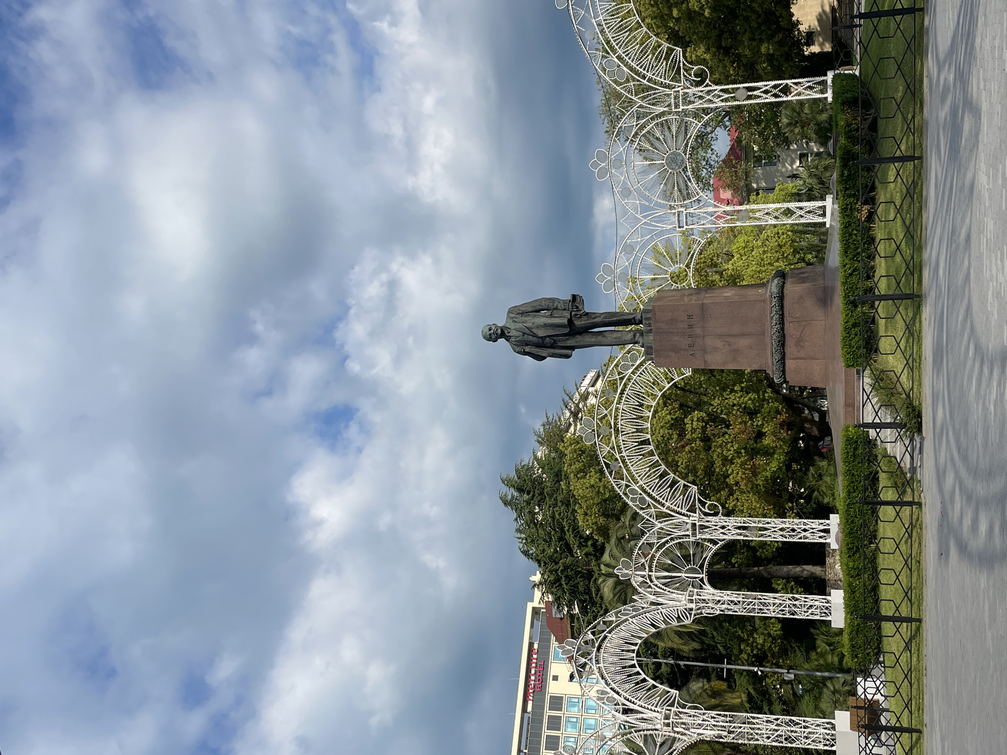 The image shows a statue of Vladimir Lenin in Sochi, Russia. The statue stands on a stone pedestal with the name "Lenin" inscribed in Russian. The statue is framed by decorative white arches. The background includes trees, a building with the word "Mercure Hotel" in red, and a cloudy sky. A black metal fence separates the statue and greenery from the pavement.
