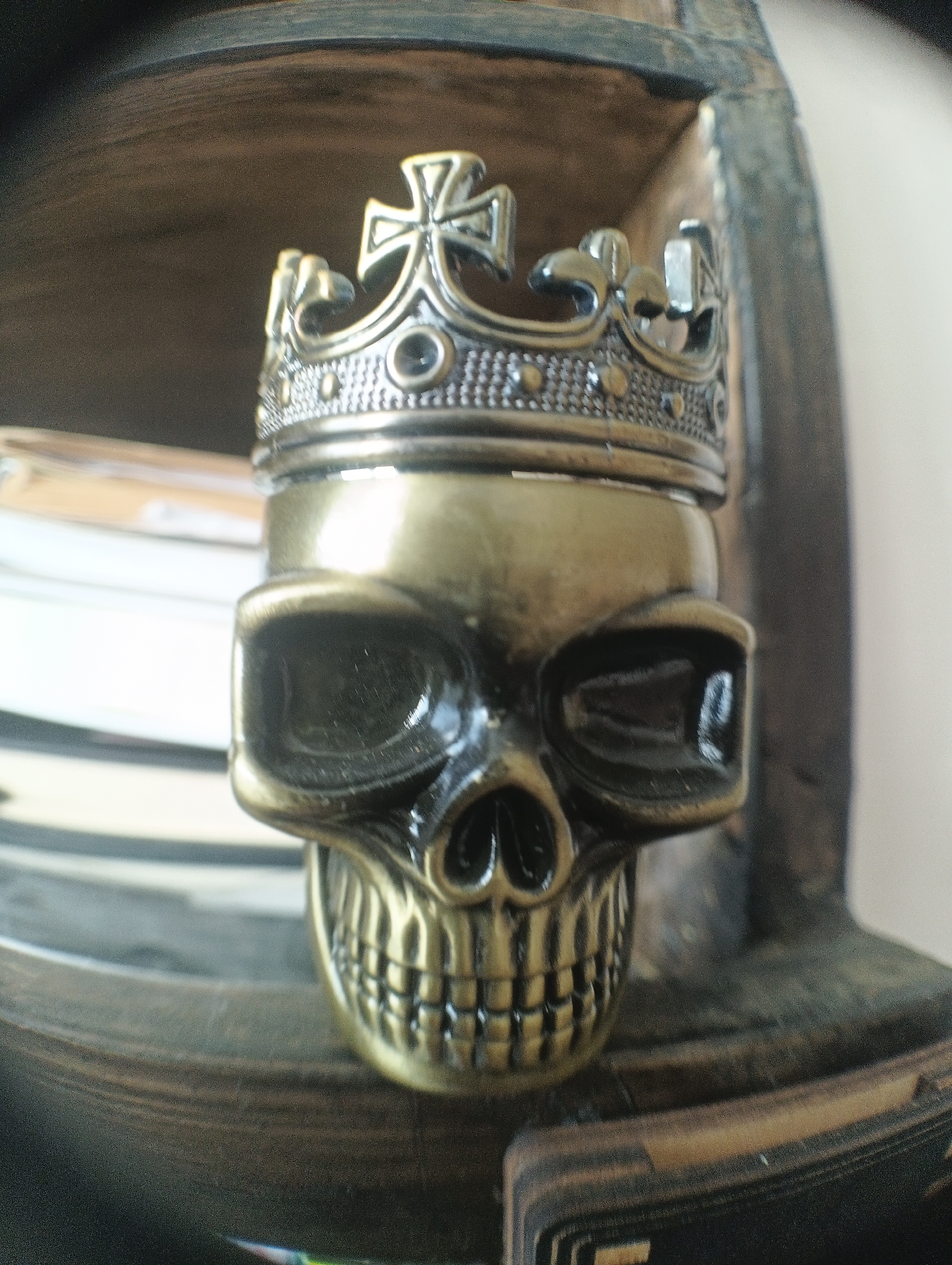 The image presents a close-up of a metallic skull figurine wearing a crown, positioned within a dark, possibly wooden, interior space, such as a shelf or a box. The figurine is crafted from what appears to be brass or an antique gold-colored metal, exhibiting a somewhat reflective surface.
The skull itself features distinct, darkened eye sockets, a prominent nose cavity, and a detailed set of teeth. Adorning its cranium is an intricately designed crown, also metallic, which adds a regal or gothic theme to the object. The crown displays several ornamental elements, including a prominent cross at its highest point, surrounded by other decorative motifs and possibly small studs along its band. The overall impression is one of detailed craftsmanship.
In the blurred background behind the skull, a stack of light-colored objects, likely books or papers, can be vaguely discerned. The immediate setting around the skull consists of dark, textured surfaces, suggesting wood with visible grain or a similar material. The lighting creates highlights on the metallic surface of the skull and crown, indicating an indoor environment with ambient light.
No activity or event is taking place; the image is a still life focusing on the decorative object. There is no visible text within the frame. The photograph features a shallow depth of field, keeping the skull and crown in sharp focus while blurring the foreground and background elements. The location context for this scene is Karnobat, Bulgaria.