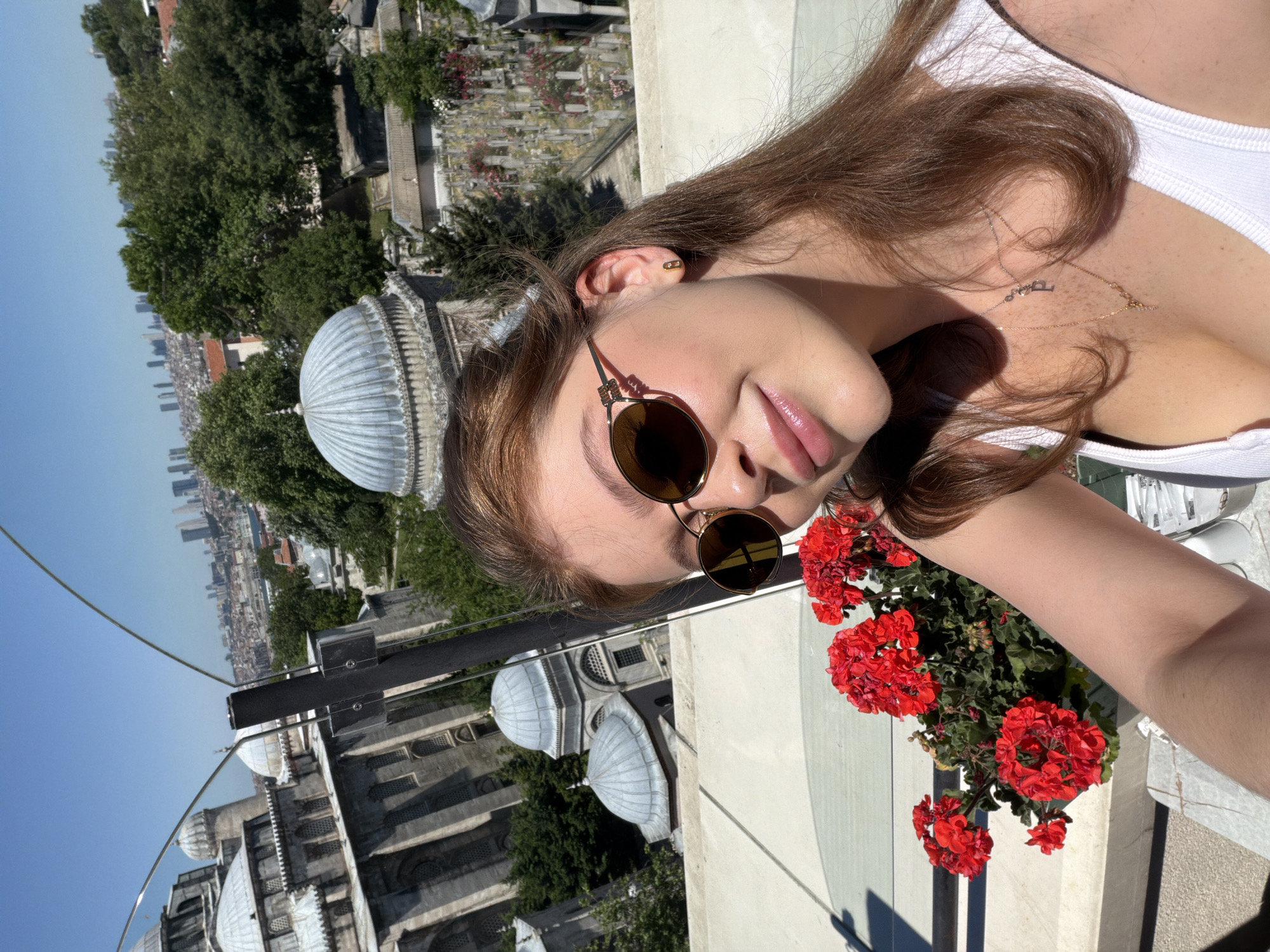 The image is a selfie of a woman with brown hair and sunglasses, wearing a white top, and smiling slightly at the camera. Behind her is a cityscape, including domes and other architectural features characteristic of Istanbul, Turkey. Specifically, the Suleymaniye Mosque is visible, indicating that the photograph was taken from or near the rooftop area of a building with a clear view towards the mosque and the broader cityscape. There is also greenery, including trees, and a flower box with bright red flowers is in the foreground. The weather appears sunny.