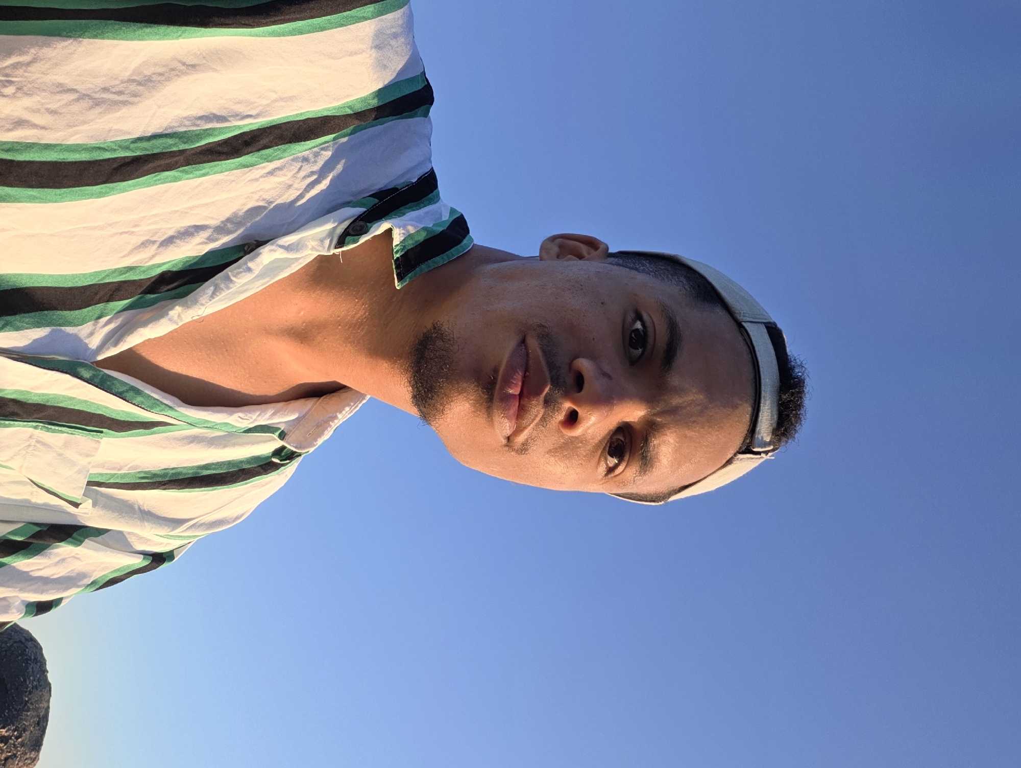 A head and shoulders portrait of a man with dark curly hair and a light-colored baseball cap is captured against a clear blue sky. He is wearing a vertically striped short-sleeved shirt with white, black, and green stripes. The man has a light brown complexion, a small goatee, and is looking directly at the camera with a neutral expression. The lighting suggests it is either early morning or late afternoon. A dark, textured object, possibly a rock, is visible in the lower right corner of the frame. The setting appears to be outdoors, with no discernible urban structures or Varna-specific landmarks visible in the image.