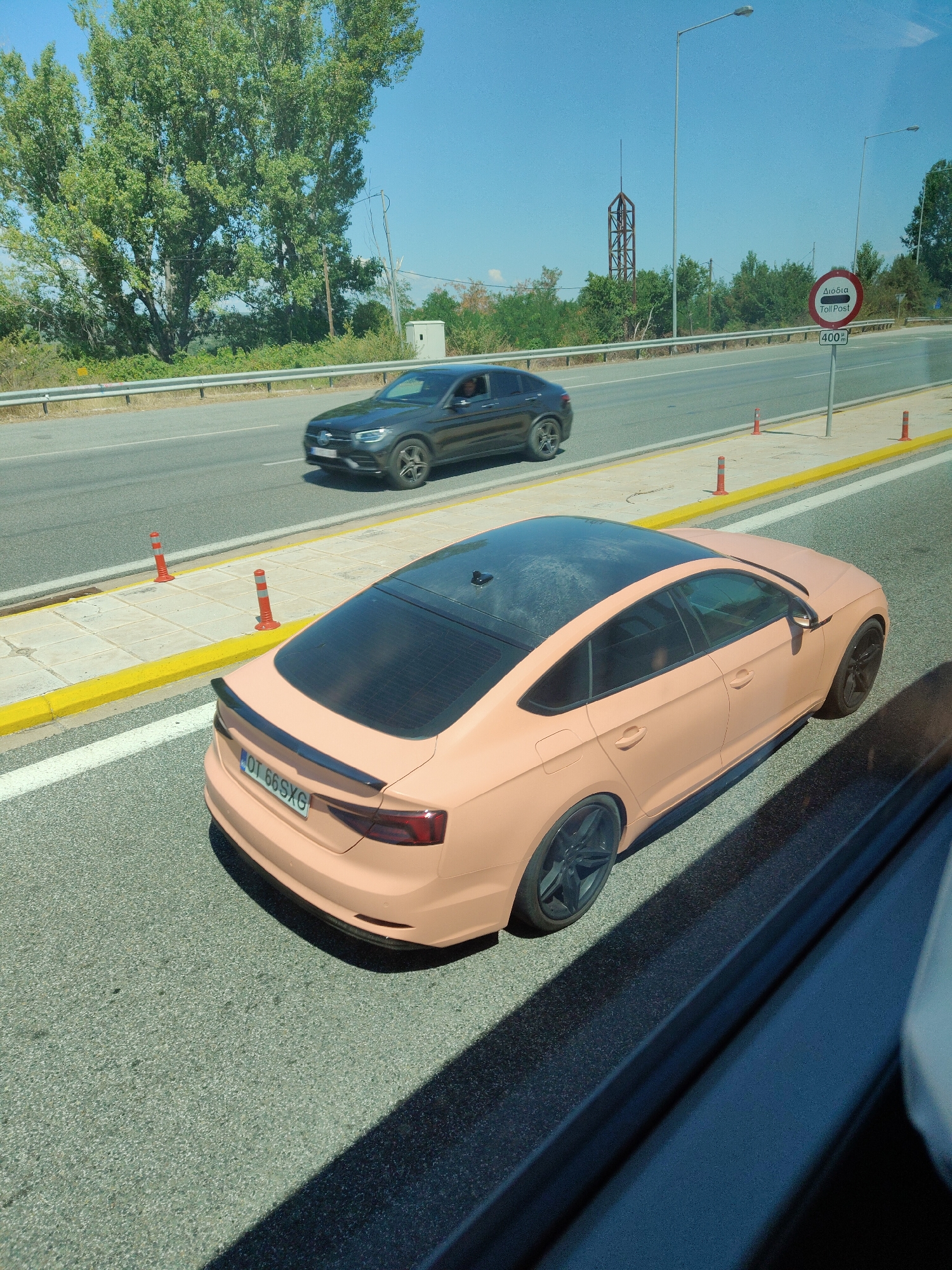 The image depicts a street scene in Δημοτική Ενότητα Προμαχώνος, Greece, under a clear blue sky. In the foreground, a peach-colored Audi A5 Sportback with a black spoiler and tinted windows is parked on the right side of the road. Its license plate reads "OT 66SXG". To the left of the Audi, a dark gray Mercedes-Benz SUV is driving on the road. Orange traffic cones are visible along the side of the road. In the background, a sign indicating a "Toll Post" with "400 m" is posted. Further in the background, there are trees, a metallic structure, and streetlights. The scene suggests a toll road environment with active traffic.