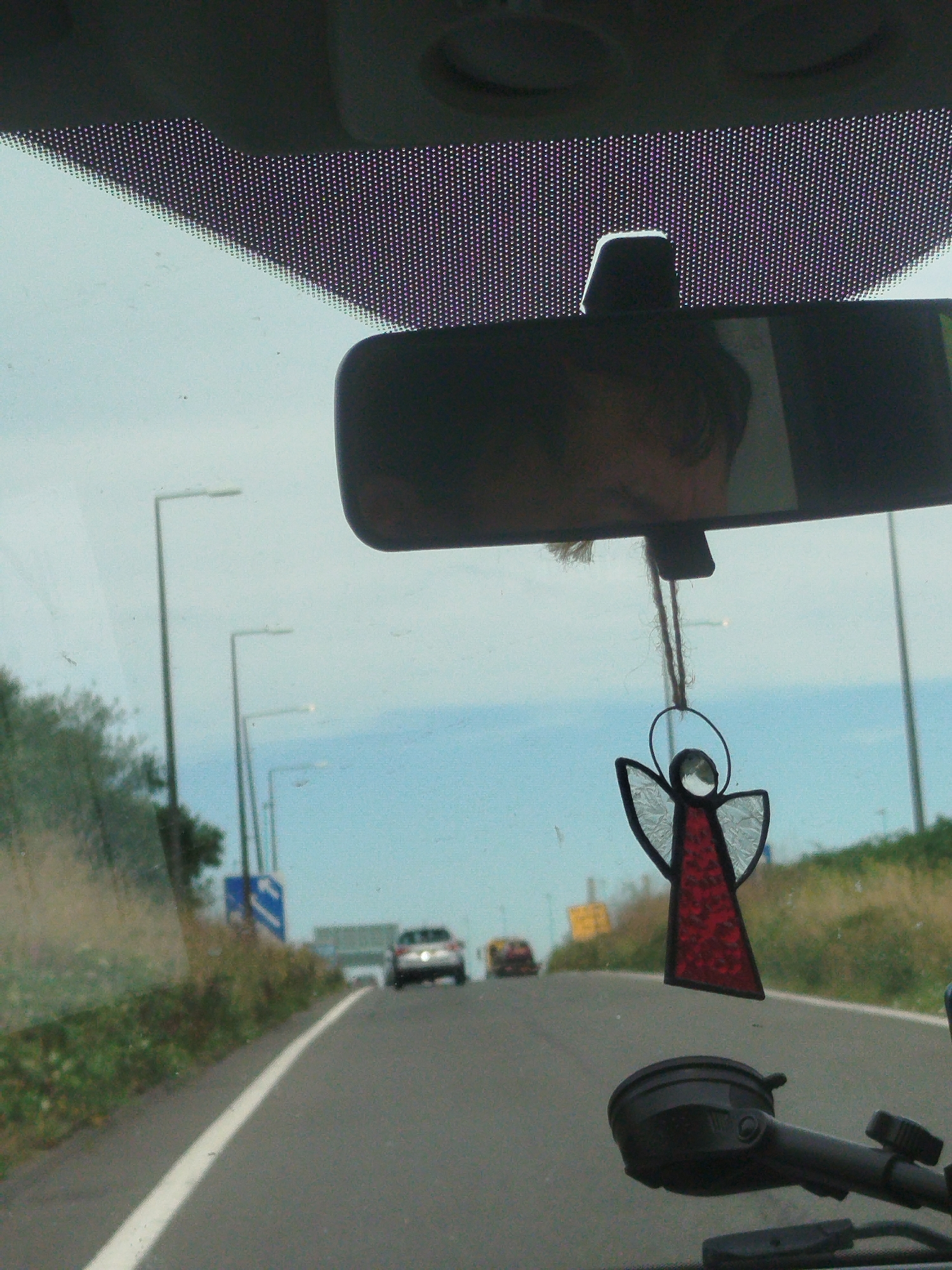 The image shows a view from inside a vehicle traveling on a road. A rearview mirror is visible at the top of the frame, reflecting a person's face. A stained-glass angel ornament hangs from the mirror. The road ahead has a white line marking the lane. Streetlights are visible on the left side of the road, and a blue road sign with a white arrow is also present. Further down the road, a car and a bus are visible. The environment appears to be a road in a semi-rural or suburban area, possibly near the coast, given the clear sky and open landscape. The image suggests movement, as it is taken from a moving vehicle. The location is identified as Severn Beach, United Kingdom.