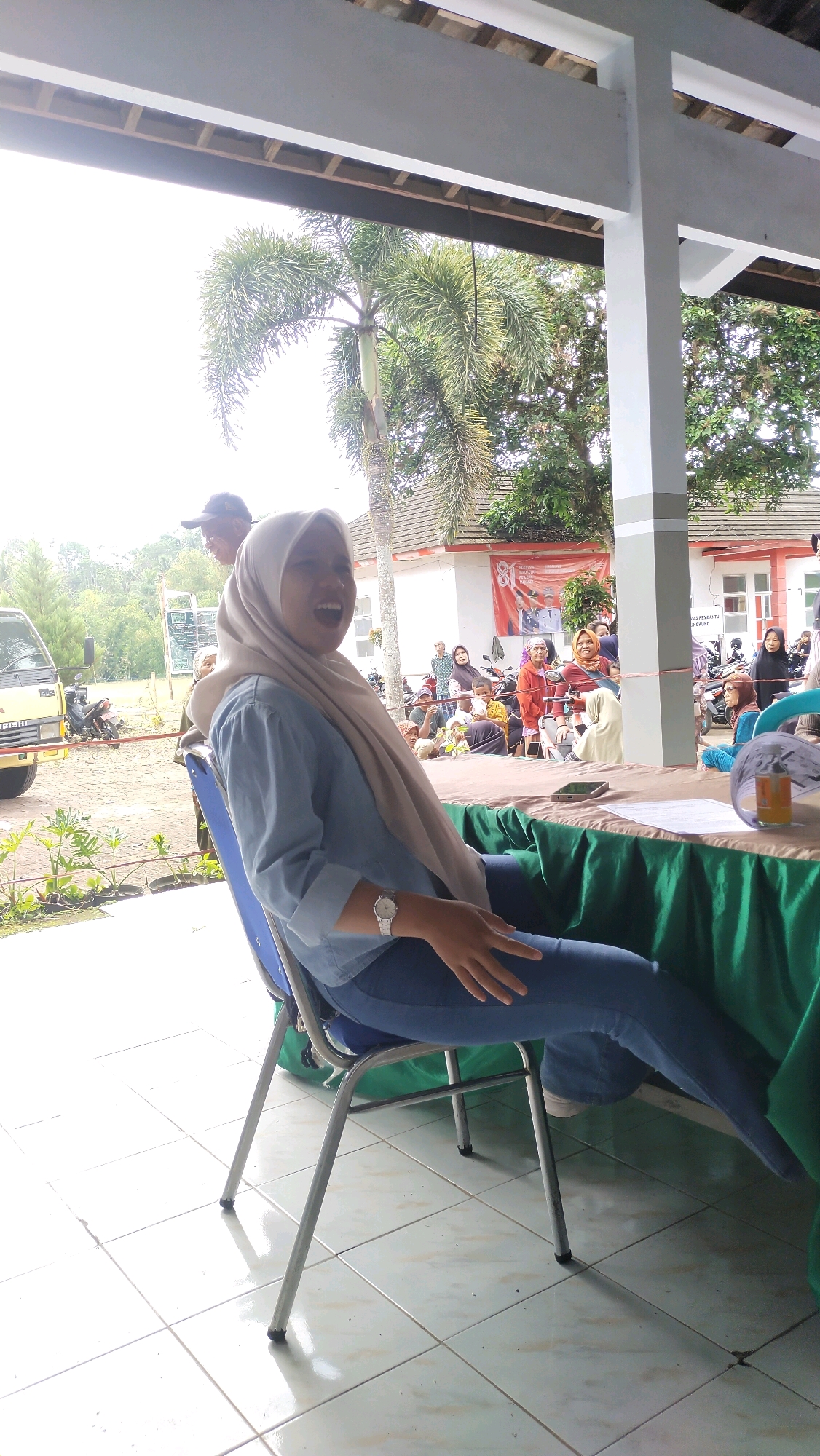 A medium-shot, eye-level outdoor scene shows a young Indonesian woman wearing a light blue long-sleeved shirt, jeans, and a light brown hijab, seated on a blue chair. She is leaning back and laughing with her mouth open, her right hand resting on her thigh and her left arm bent at the elbow, with her hand reaching towards her left side. A silver wristwatch is visible on her left wrist. Her legs are extended forward, and she is looking towards the right side of the frame.
Behind her, a table is covered with a green cloth, and on it are papers and a bottle of liquid. A dark-colored smartphone lies on the table. Further in the background, a group of people are gathered, some sitting and some standing, near motorcycles. A red banner with Indonesian text is displayed on a building. In the distance, palm trees and other greenery are visible. The ground is tiled with light-colored squares. A yellow truck is parked to the left of the frame. The scene appears to be in a public area in Klungkung, Indonesia, possibly during an outdoor event or gathering.
