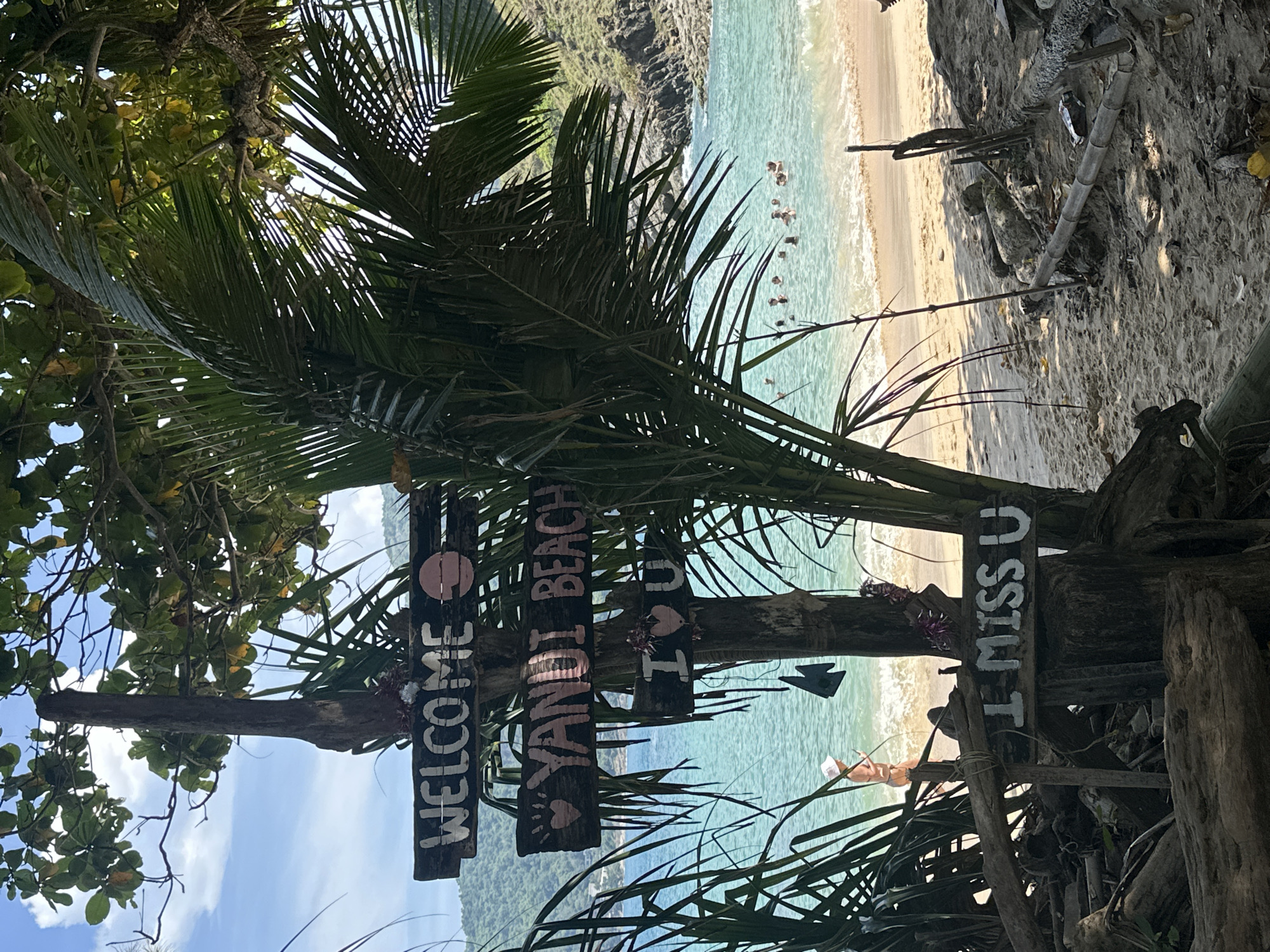 A wooden signpost with three wooden planks is situated on a sandy beach in Rawai, Thailand. The top plank reads "WELCOME" in white lettering, with a pink circle to the right. The middle plank says "YANDI BEACH" in white lettering and is adorned with two pink hearts on the left. The bottom plank displays "I MISS U" in white lettering, also with two pink hearts on the left, slightly above the text. Behind the signpost, clear blue water with gentle waves laps onto a sandy shore. Several people are visible swimming and enjoying the water in the distance. Lush green vegetation, including palm trees, frames the scene. The overall environment suggests a tropical beach setting on a bright, sunny day.