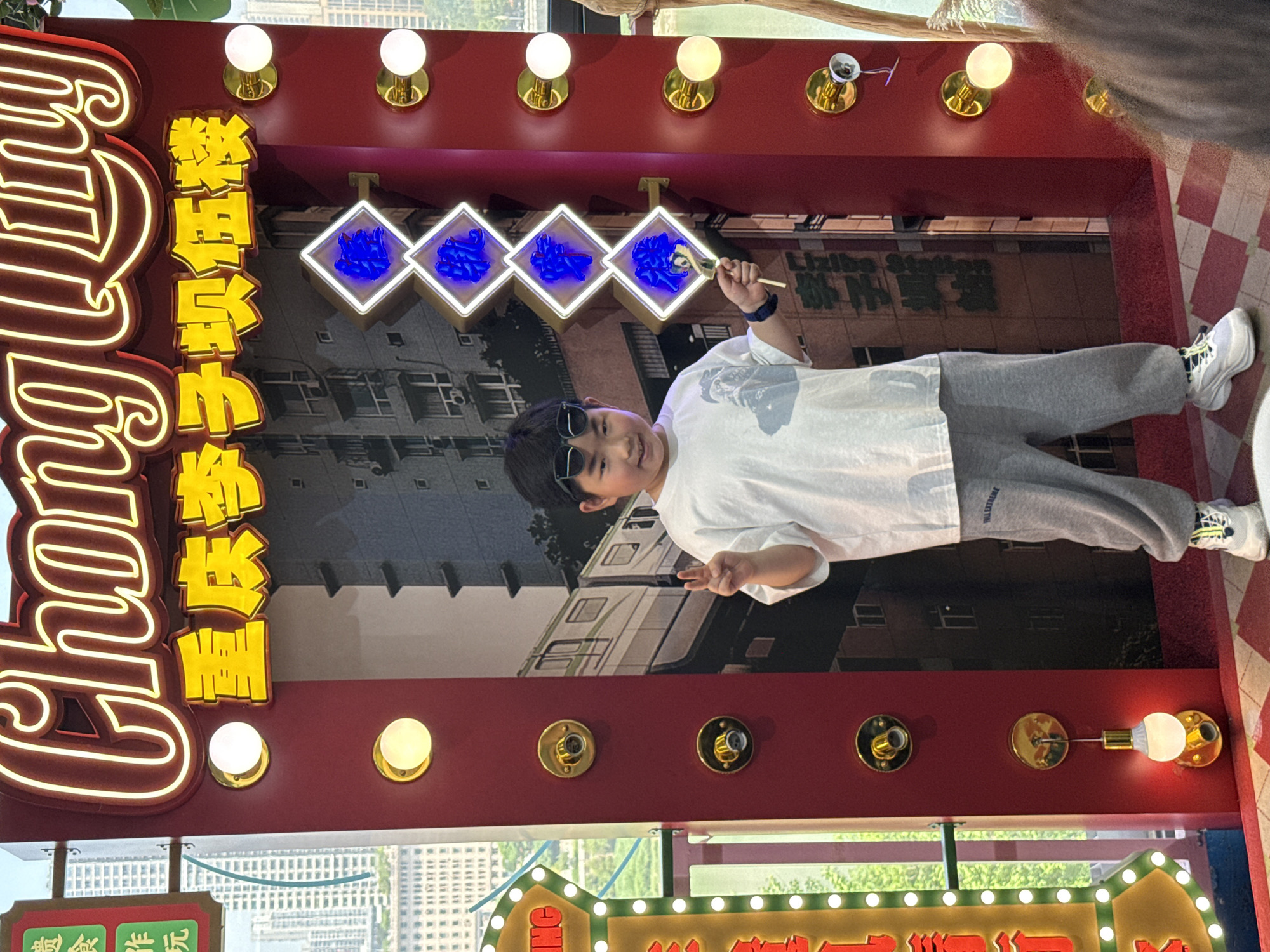 The image captures a young boy posing playfully within a brightly lit, red-framed display area, likely an interactive exhibit or photo booth. The scene is set indoors or under a covered structure, with natural light visible in the background, suggesting it is daytime.
The main subject is a boy with dark hair and black sunglasses pushed up onto his head. He is wearing a white T-shirt with a graphic print, grey sweatpants with "EXTREME" visible on the left leg, and white and yellow sneakers. He has a cheerful expression, smiling directly at the camera, and holds a paintbrush-like object with chopsticks in his left hand, gesturing towards the backdrop.
The display features a prominent red frame with golden light fixtures, some illuminated, lining the top and bottom. On the left side of the frame, large white neon letters spell out "Chongqing" (partially visible), and yellow neon Chinese characters arranged vertically read "繁华都市" (Fánhuá Dūshì), which translates to "Prosperous City." The backdrop behind the boy depicts an urban landscape with buildings and a train, typical of Chongqing. Text on the backdrop includes "Liziba Station" and "鹅岭贰厂" (Élíng Èrchǎng), known landmarks in Chongqing's Yuzhong District. Four diamond-shaped frames with blue illuminated elements are arranged horizontally across the top of the backdrop. Below the main display, a green sign adorned with white lights reads "好吃好玩" (Hǎochī Hǎowán), meaning "Delicious and Fun." The overall atmosphere is vibrant and inviting.