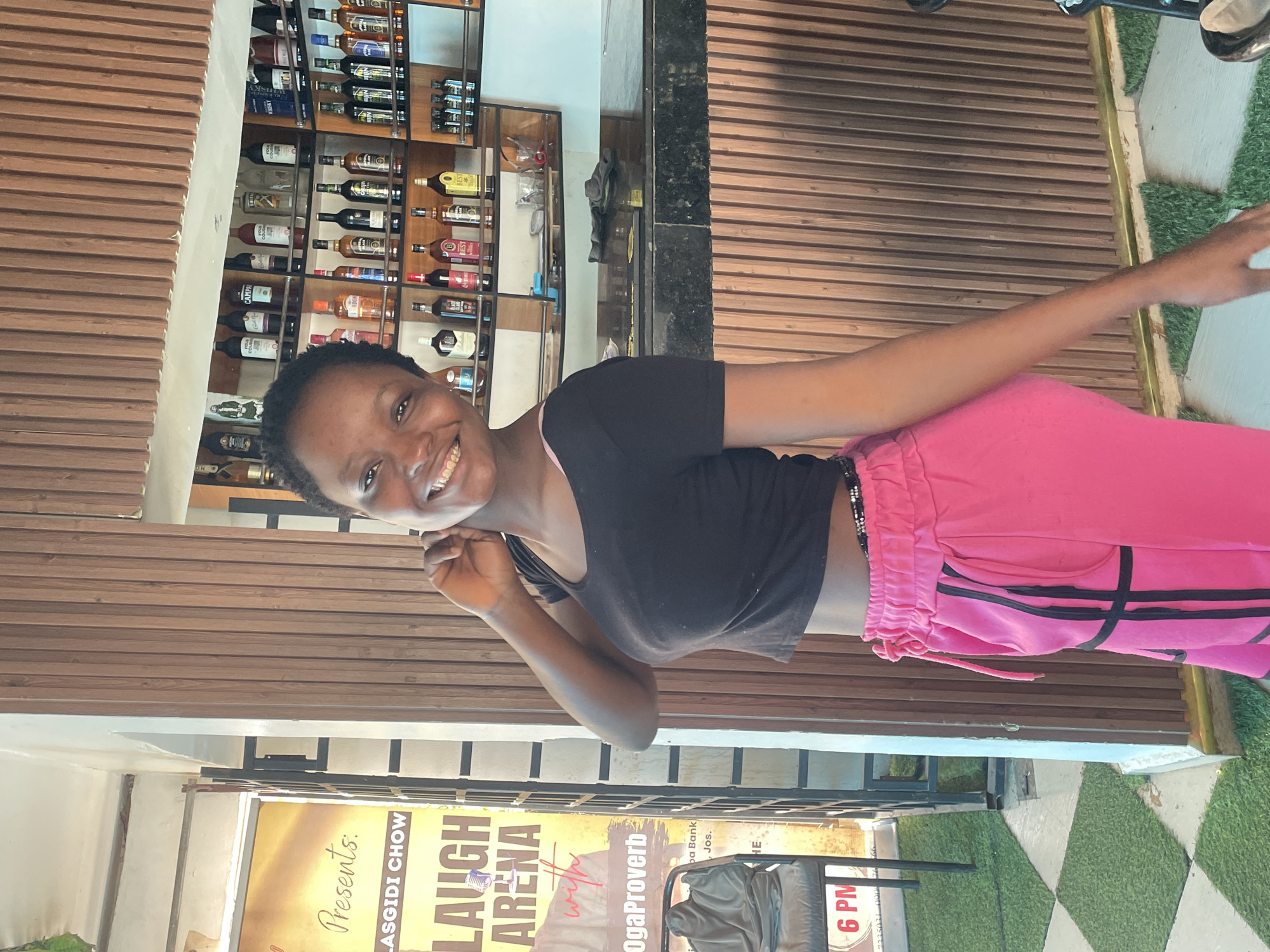 In Jos, Nigeria, a young woman with dark skin and short hair smiles at the camera, her right hand near her ear. She is wearing a black crop top and bright pink sweatpants with black stripes. She stands in front of a wooden wall behind a bar, where bottles of liquor are displayed on shelves. To her left, a yellow poster advertises "LAUGH ARENA" with the words "Presents:" and "LASGIDI CHOW" and "OgaProverb". A chair with a grey cloth is visible next to the poster. The floor is a checkered pattern of grey and artificial grass.