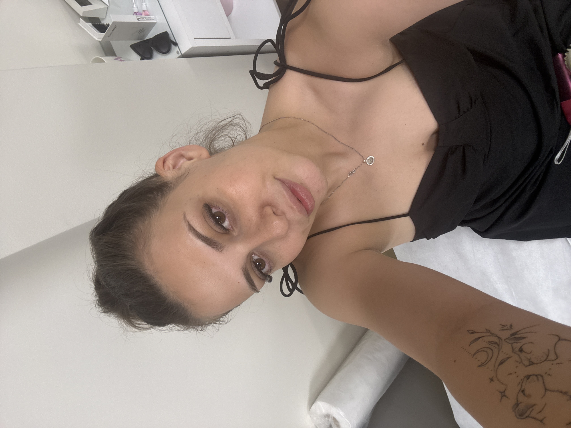 The image shows a woman with dark hair tied up, wearing a black slip dress and a silver necklace. She has a tattoo on her left arm. She is indoors, with a white wall and a white roll of fabric in the background. A white dresser with some products and sunglasses on top is visible to the right. The overall lighting is bright, suggesting an indoor setting with artificial light. There is no indication of the specific location in Plovdiv, Bulgaria.