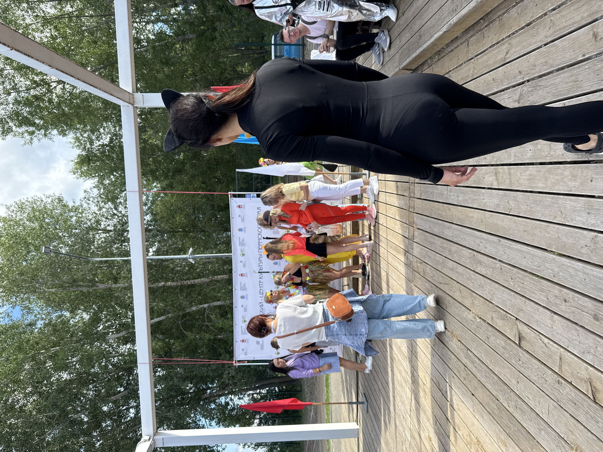 The image depicts an outdoor event, likely a festival or competition, taking place on a wooden boardwalk under a bright sky with scattered clouds. The setting appears to be near a body of water, indicated by the presence of trees lining the background and a distant blue structure that could be a vehicle or building. In the foreground, a person wearing a black full-body suit and cat ears walks away from the camera. To the left, a group of people are gathered near a white banner with Russian text, suggesting an official presence or sponsorship. Several individuals are dressed in colorful, festive attire, including hula skirts, leis, and bright jumpsuits. A red flag on a pole stands to the left of the group. The overall atmosphere suggests a lively and informal gathering. The banner's text, which appears to read "ЧАЙКОВСКИЙ – ЦЕНТР КУЛЬТУРЫ ПЕРМСКОГО КРАЯ" (CHAİKOVSKY – CULTURE CENTER OF PERM KRAY), firmly places the event in Chaikovsky, Russia.