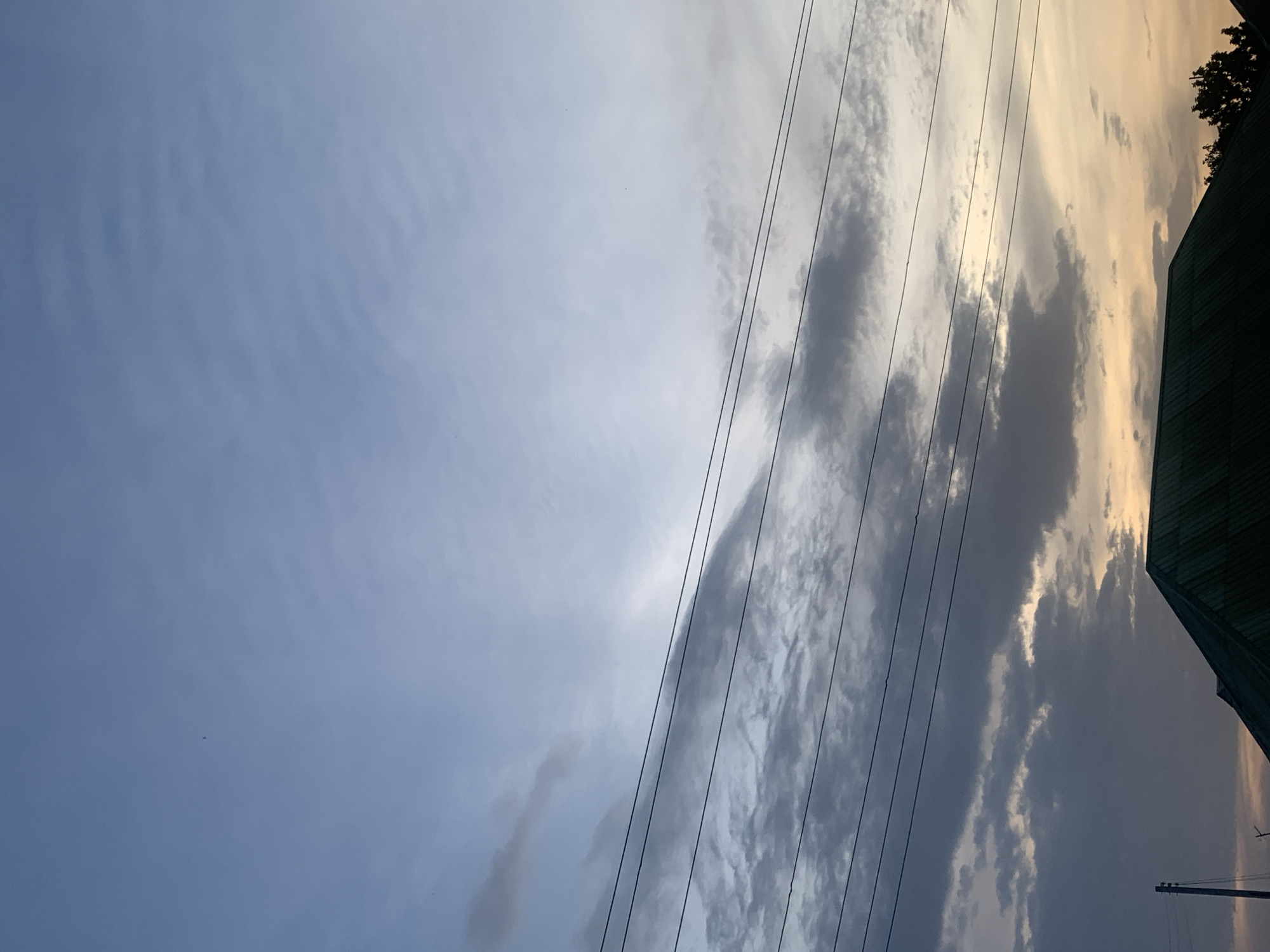The image displays a sky scene, likely captured during late afternoon or dusk. When viewed with the horizon oriented correctly, the left side of the frame shows a clear, light blue sky with faint, wispy cloud patterns. Towards the center and right, the sky transitions, becoming filled with more substantial cloud cover. These clouds are a mix of dark gray and brighter, yellowish-orange hues, suggesting the warm light of a setting sun filtering through or reflecting off them.
Several parallel power lines run diagonally across the middle and right portions of the frame. In the bottom right corner, the dark, angled roof of a building, possibly a house or a similar structure, is visible. Above the roof, the dark silhouette of tree foliage is discernible against the lighter sky. A utility pole is also faintly visible in the far bottom right. There are no people or specific activities taking place, and no visible text. The scene captures the interplay of natural light and clouds with man-made infrastructure in Barakin Akawo, Nigeria.