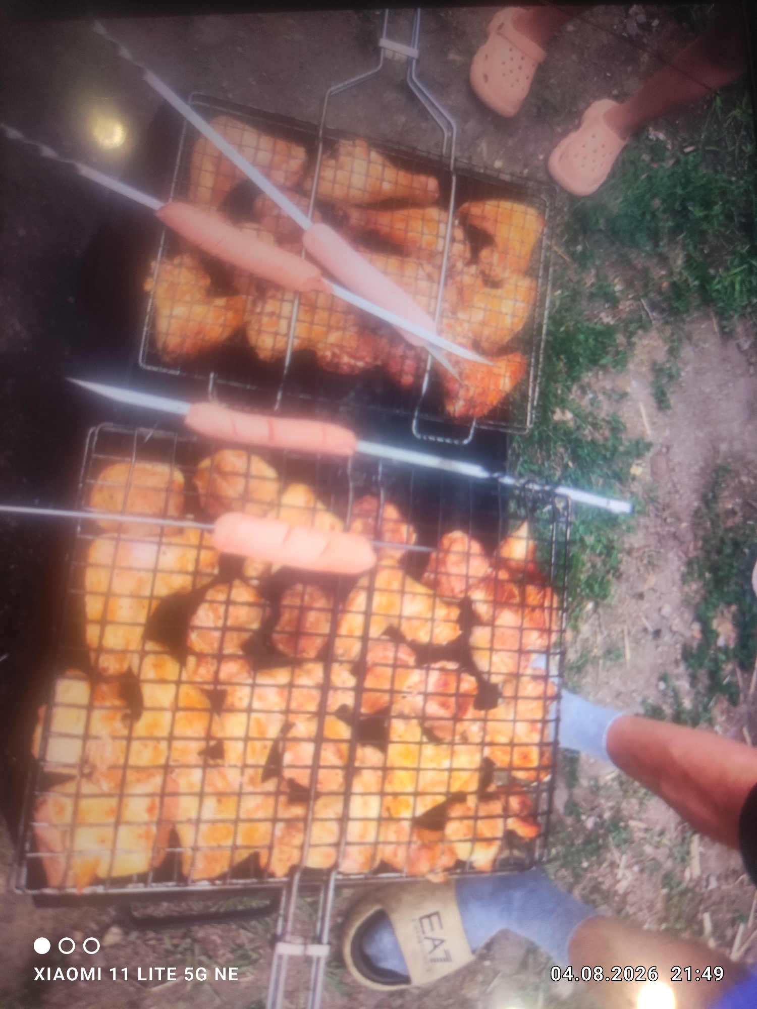 Two metal grilling baskets are positioned on the ground, filled with pieces of meat and sausages. In the upper basket, sausages are skewered and placed over pieces of chicken. The lower basket contains more pieces of meat. The scene appears to be an outdoor setting with green grass and dirt. In the upper right of the image, two people's feet are visible, wearing light-colored clogs. In the lower right, a person's legs and feet are visible, one wearing a light-colored sock and the other a sandal with the text "EAT" visible. A timestamp in the lower right corner reads "04.08.2026 21:49", and a watermark in the lower left reads "XIAOMI 11 LITE 5G NE". The image is taken from a high angle looking down. The location is specified as Kara-Chungur, Kyrgyzstan.