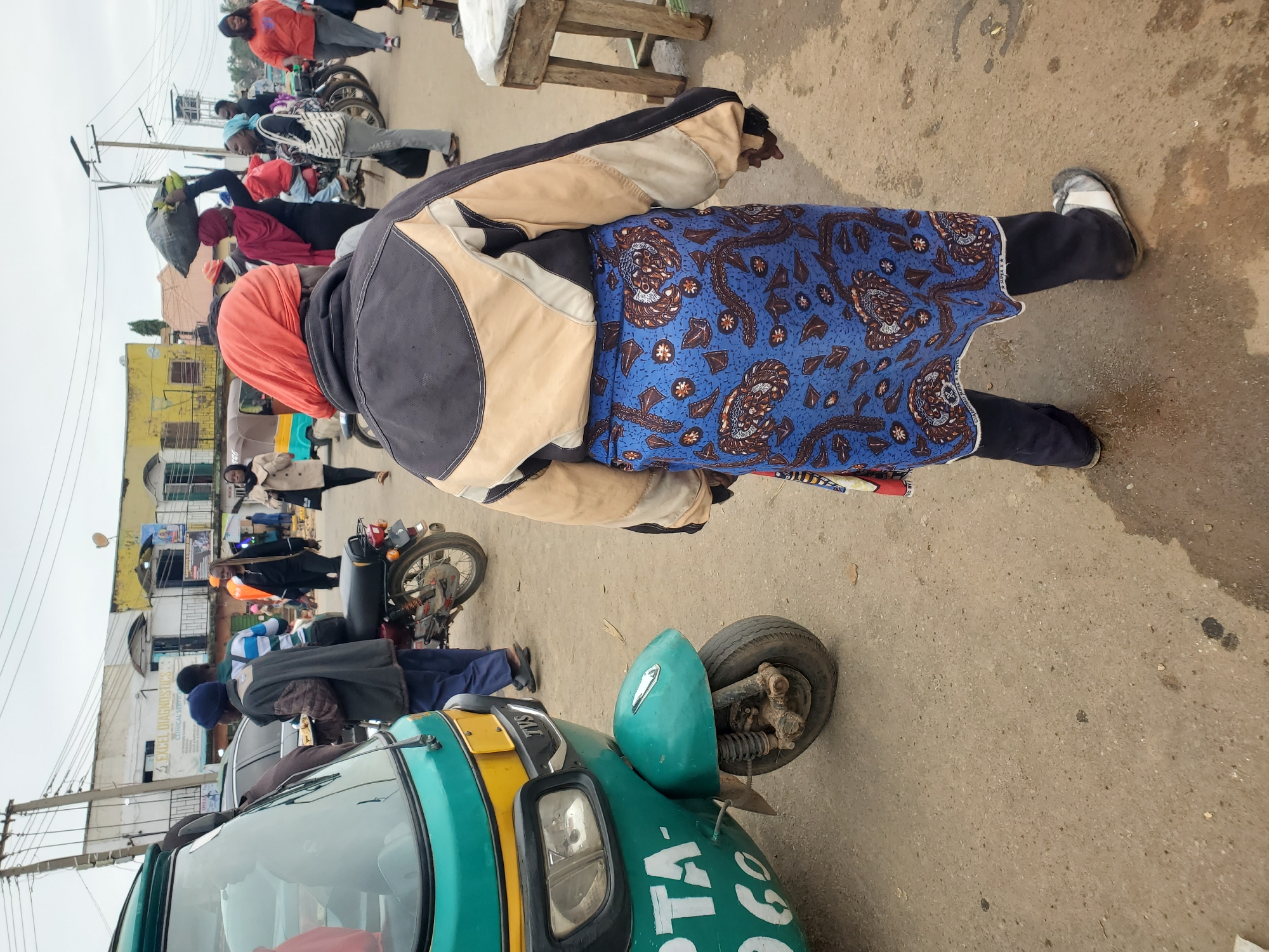 In Gwong, Nigeria, a person wearing a blue patterned skirt, dark pants, and a jacket with beige and black accents stands in a dusty, outdoor marketplace. The individual has an orange headscarf and faces away from the camera. A turquoise and yellow three-wheeled vehicle, possibly an auto-rickshaw, is parked to the left, with partial text reading "PTA-960" visible on its side. Several other people are in the background, some carrying loads and others near motorcycles and buildings. The environment appears to be a busy street with overcast skies.