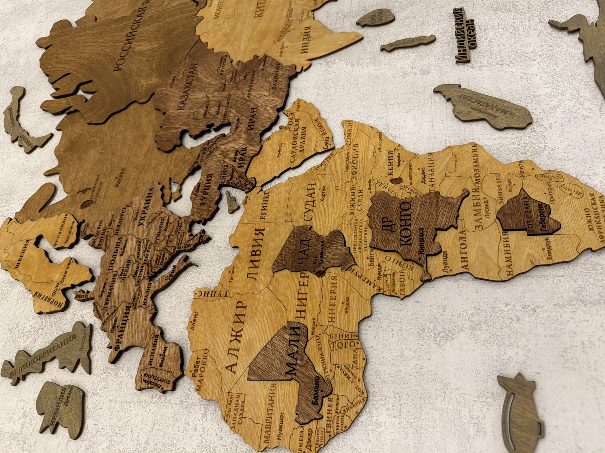 The image displays a wooden world map with country names and some cities labeled in Russian. The map is mounted on a textured, light-colored surface. Several countries and regions are represented by cut-out wooden pieces. Notable landmasses include Europe, Africa, and parts of Asia. Individual country names visible include "РОССИЙСКАЯ Ф" (Russian Federation), "КАЗАХСТАН" (Kazakhstan), "ИРАК" (Iraq), "ИРАН" (Iran), "ИНДИЯ" (India), "КИТА" (China), "ТУРЦИЯ" (Turkey), "ЕГИПЕТ" (Egypt), "СУДАН" (Sudan), "ЛІВІЯ" (Libya), "ТУНИС" (Tunisia), "АЛЖИР" (Algeria), "МАЛІ" (Mali), "НИГЕР" (Niger), "ЧАД" (Chad), "НИГЕРИЯ" (Nigeria), "КОНГО" (Congo), "АНГОЛА" (Angola), "ЗАМБИЯ/МОЗАМБИК" (Zambia/Mozambique), "НАМИБИЯ" (Namibia), "БОТСВАНА" (Botswana), "МАДАГАСКАР" (Madagascar), "ФРАНЦИЯ" (France), "ИСПАНИЯ" (Spain), "ИТАЛИЯ" (Italy), "ПОЛЬША" (Poland), "ГЕРМАНИЯ" (Germany), "ШВЕЦИЯ" (Sweden), and "НОРВЕГИЯ" (Norway). Some cities are labeled, such as "МОСКВА" (Moscow) and "ДАКАР" (Dakar). Additionally, there are small wooden cut-outs shaped like airplanes and an airship scattered around the map. There is no indication of the city of Semiluki, Russia, in the provided image.