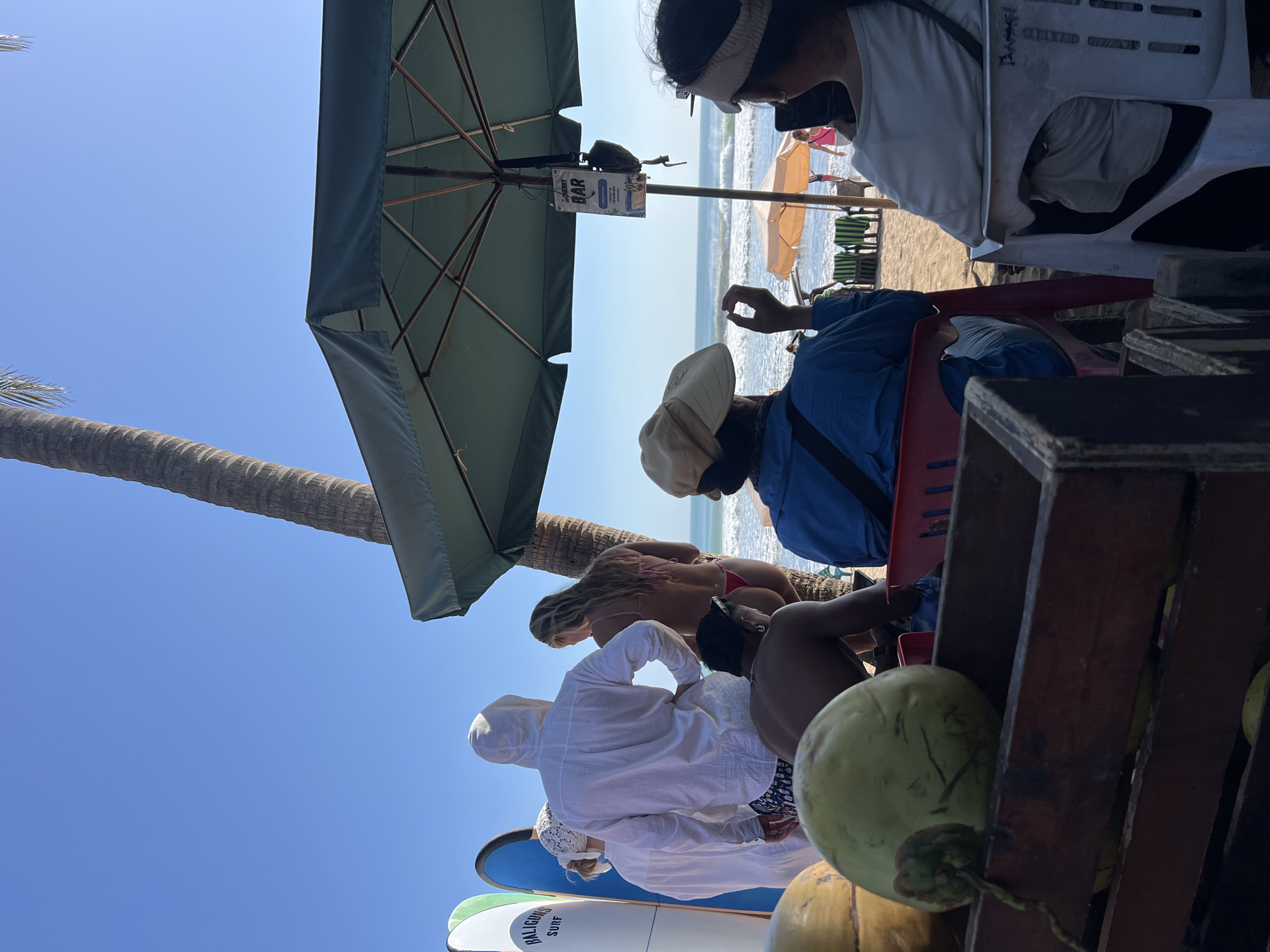 A group of people are gathered on a beach in Kuta, Indonesia, near surfboards and under a large umbrella. In the foreground, a wooden crate holds coconuts. A woman wearing a visor and a woman in a blue jacket with a hat are seated, facing away from the viewer, looking towards the ocean. Behind them, a man wearing a white hooded shirt and patterned shorts stands with his back to the camera. A woman in a pink bikini is also present, with her back to the camera. Surfboards are lined up to the left of the frame. The ocean with waves is visible in the background, under a clear blue sky. A sign for "CHERRY BAR" hangs from the umbrella.