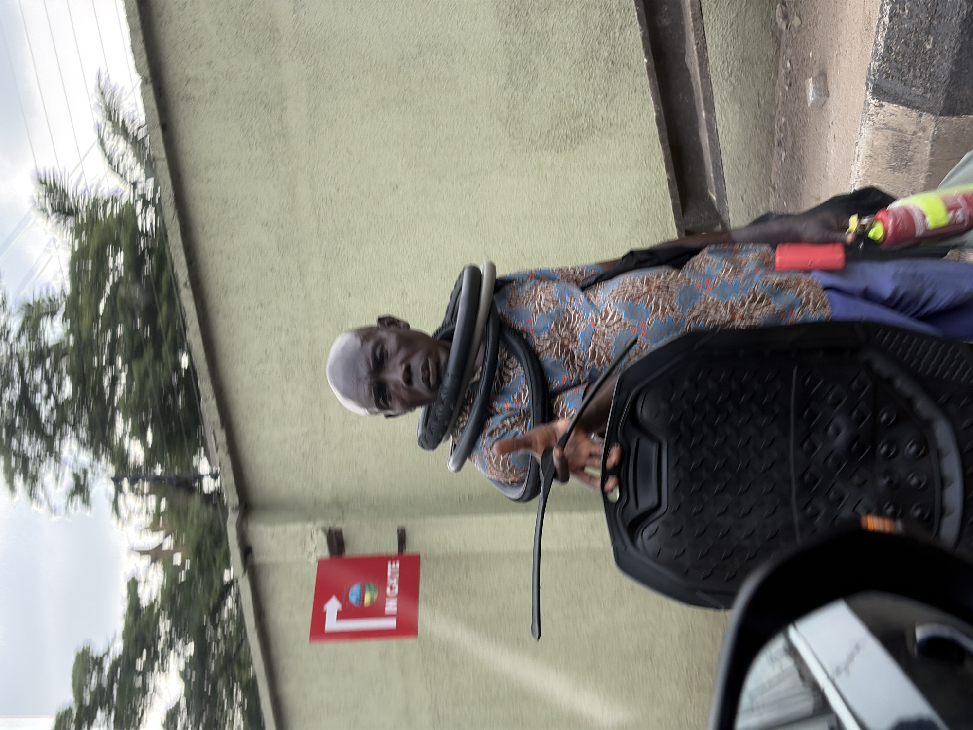 A partially visible elderly African man stands near a light yellow wall with a red sign that reads "IN GATE" next to a directional arrow. The man has a bald head with a fringe of white hair and several black hoses are coiled around his neck. He is wearing a patterned blue shirt. He appears to be on a street, with a black car floor mat and a portion of a car mirror visible in the foreground. Trees and a cloudy sky are visible on the left, suggesting the scene is outdoors. The sign and context point to this being in Lagos, Nigeria. The man's purpose and the specifics of his interaction with the vehicle are not discernible from the image.