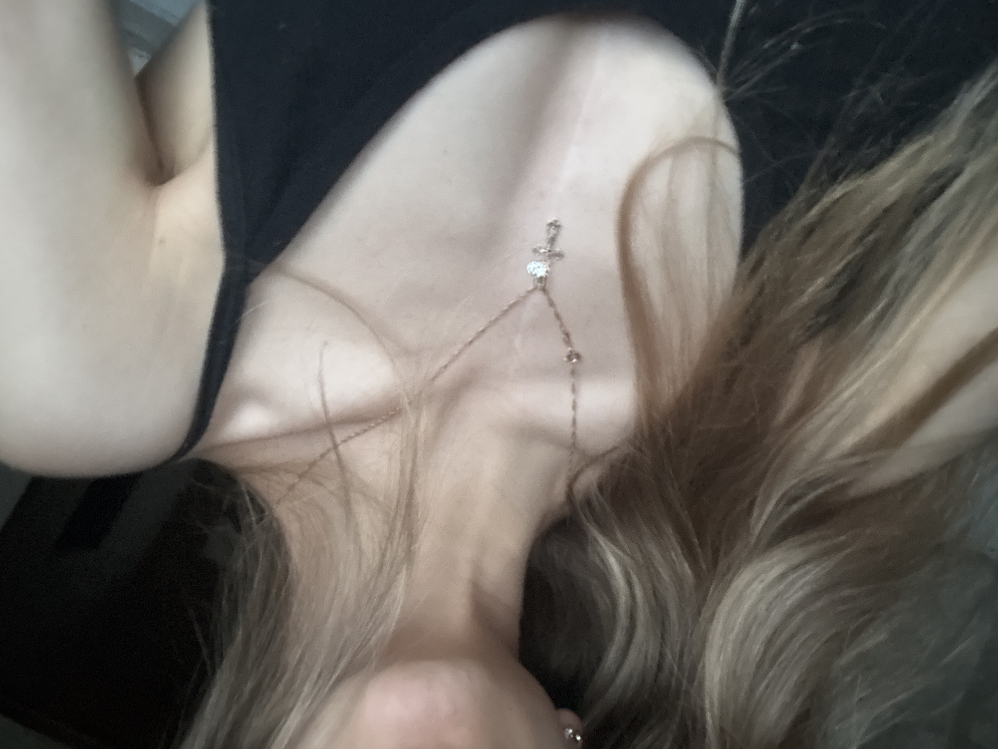 The media file presents a close-up, high-angle view of a person, likely female, captured from their chin down to the upper chest. The subject is wearing a black top or dress, visible on the upper left and right sides of the frame. A delicate gold or silver-toned necklace adorns the person's neck, featuring a small cross-shaped pendant positioned centrally below the throat, and a heart-shaped stone situated slightly lower on the chain. The necklace chain extends downwards on both sides. Long, light blonde and brown hair is prominent, framing the face and flowing around the neck and shoulders, with various strands visible across the image. The person's chin and a hint of the lower lip are visible at the bottom of the frame, suggesting the person is looking upwards or has their head tilted back while lying down. This pose conveys a sense of relaxation or comfort. The background is dark and blurred, indicating an indoor environment. No specific activity beyond posing for the photograph is discernible. The time of day cannot be determined from the lighting, and there are no explicit location cues within the image itself to suggest its context in Noviy, Russia. There is no visible text.