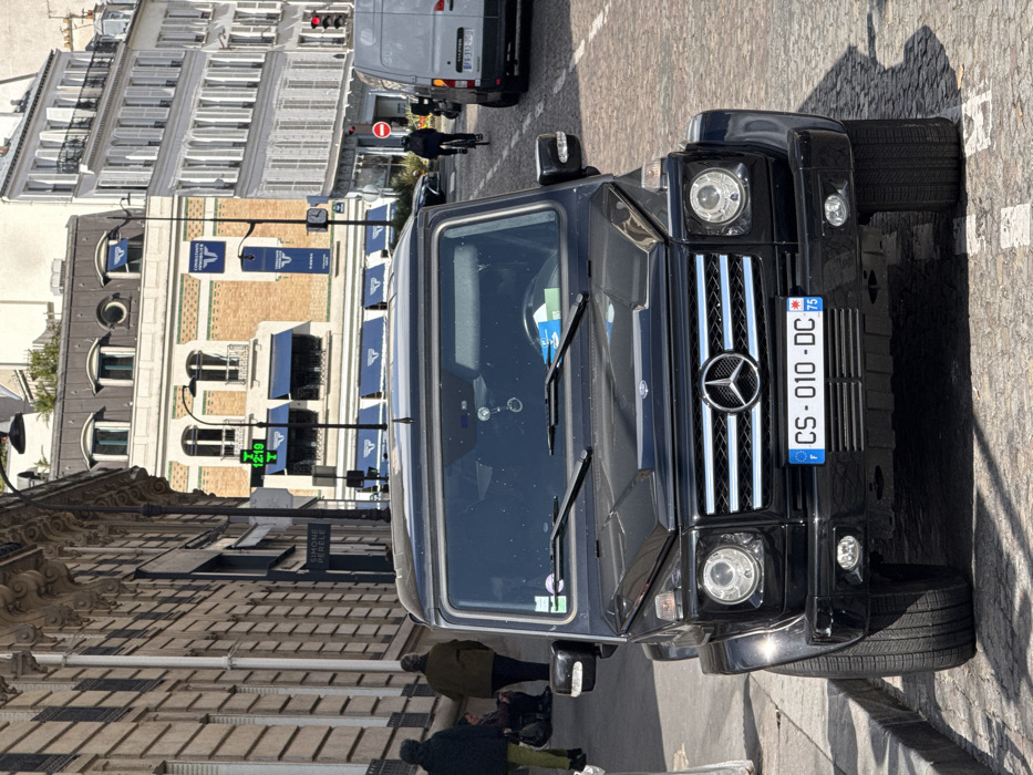 A black Mercedes-Benz G-Class SUV, bearing French license plate CS-010-DC (Paris, department 75), is parked in the foreground on a cobblestone street in Paris, France. The vehicle faces the viewer, displaying its Mercedes-Benz emblem, grille, and circular headlights.
On the left sidewalk, two male pedestrians walk past a building with a textured stone facade and a "SIMONE PÉRÈLE" sign. Further along the sidewalk, a third individual is seated. The street surface transitions from cobblestones to a slightly smoother area for the parked SUV.
In the midground, to the right of the G-Class, a male cyclist in dark clothing rides in the street, followed by a white commercial van with a visible French license plate. A "no entry" sign and a traffic light displaying red are visible further down the street. A tall street pole features a digital display showing "12:19" and "7°C".
The background is dominated by Parisian architecture. A distinctive building features a brick lower section, a lighter stone upper section, and a dark mansard roof with ornate windows. This building displays blue awnings over ground-level storefronts and vertical banners for "CONSULTANTS T". To its right, a multi-story white building with numerous windows, many fitted with closed white shutters, extends into the distance. The scene is illuminated by natural daylight, casting shadows to the right of the parked SUV.
