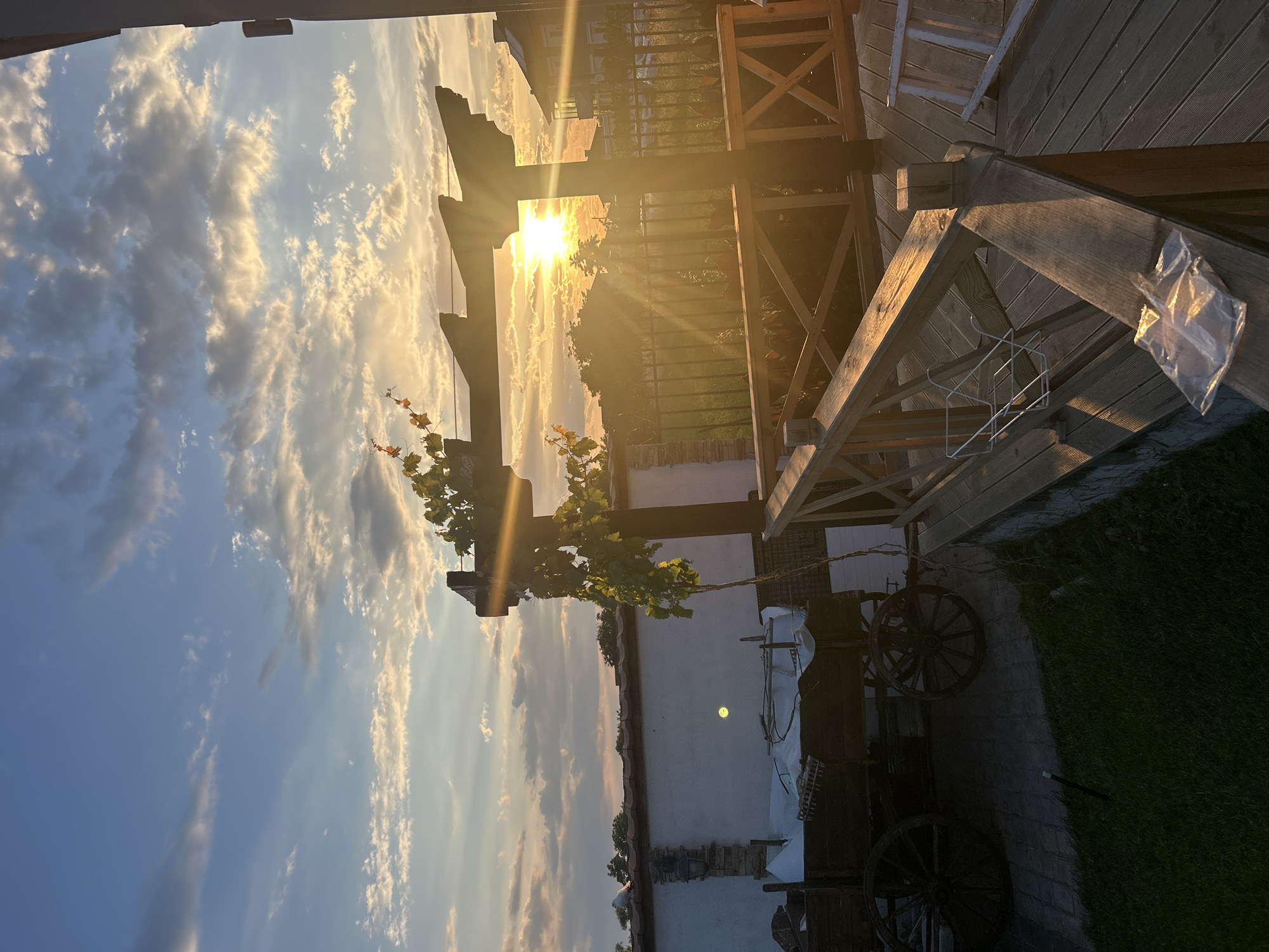 The image depicts a sunset over a rural property in Voyvodino, Bulgaria. The sky is filled with clouds, illuminated by the setting sun which casts long rays of light. In the foreground, a wooden deck with a railing is visible. A plastic bag rests on the railing. To the left, there is a white wall with a stone accent, and next to it, an old wooden cart with large wheels. A vine with orange flowers is growing on a wooden structure. Beyond the deck, a metal fence encloses a grassy area with some plants. In the background, houses and trees are silhouetted against the bright sky.