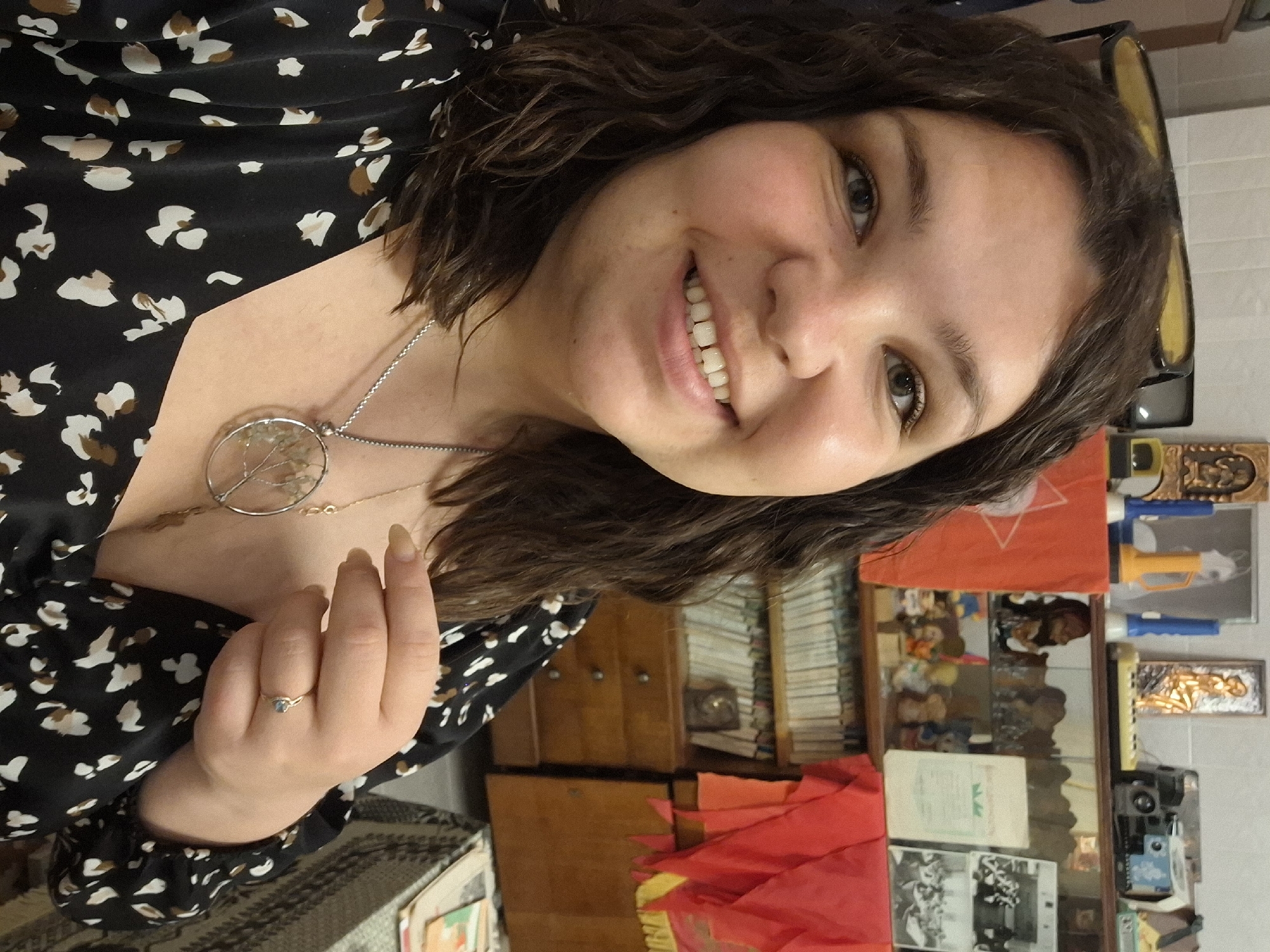 A woman with dark, wavy hair is smiling at the camera. She is wearing a black, patterned dress and a silver necklace with a circular pendant. She has a ring on her finger. Behind her is a wooden cabinet, a display case containing various objects including toys and photographs, and a stack of books. A red flag with a white design is visible in the background. While the location cannot be verified with certainty based solely on the image, the presence of Soviet-era items such as the red flag suggest a setting in a post-Soviet country, potentially Russia. I cannot verify the location of Марковский, Russia as the image provided does not include such information.