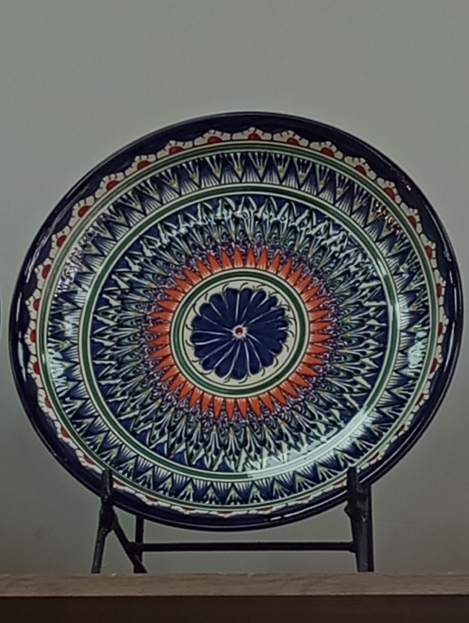 The image depicts a decorative ceramic plate displayed on a black metal stand. The plate has a circular shape and is adorned with a concentric pattern featuring predominantly blue, green, and orange colors. The central design includes a blue floral motif. The outer rim of the plate is decorated with a pattern of alternating red and white elements. The plate is placed on a shelf in front of a plain, light-colored wall. There is no evidence to suggest that the location is in Izhevsk, Russia.