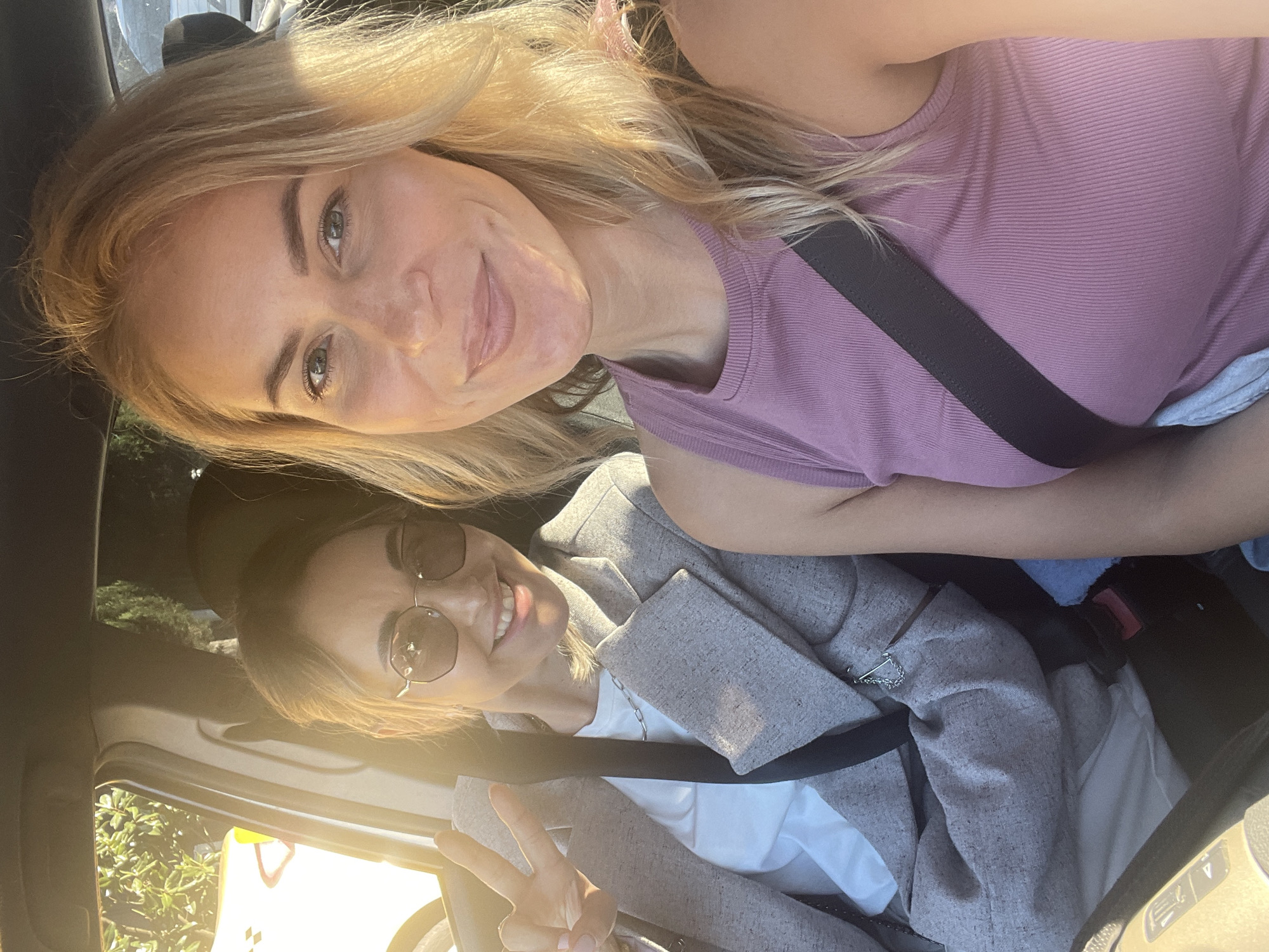 Two women are in a car. The woman in the foreground is wearing a mauve ribbed tank top and a seatbelt. The woman in the background is wearing sunglasses and a gray blazer over a white shirt, also with a seatbelt. She is making a peace sign with her hand. The location is likely Sochi, Russia. There is sunlight and foliage visible outside the car window.