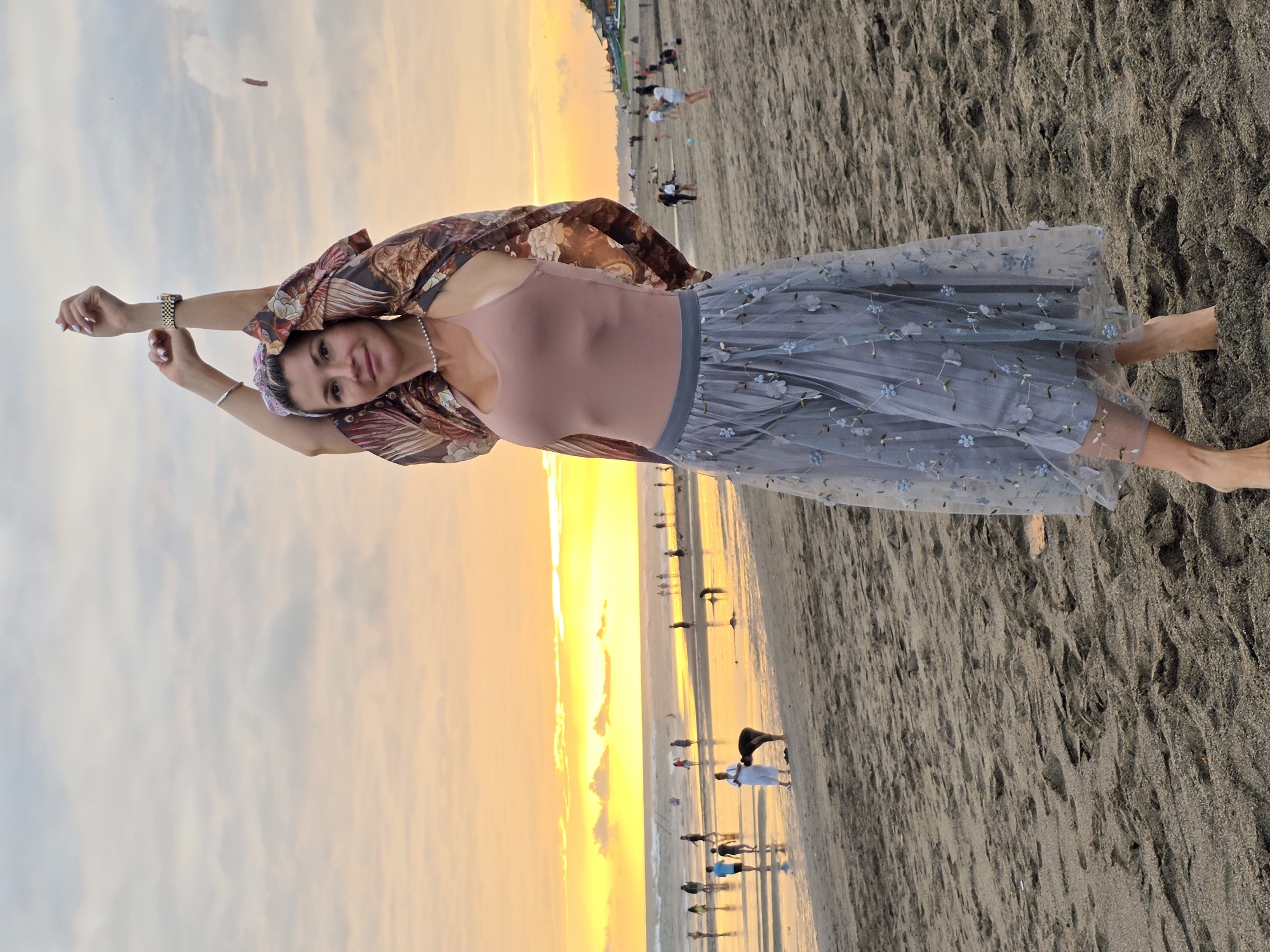 A woman stands on a sandy beach at sunset in North Kuta, Indonesia. The sky is filled with warm hues of orange and yellow, with scattered clouds. The woman is wearing a pink tank top, a sheer grey skirt with floral embroidery, and a patterned kimono-style cover-up. Her arms are raised above her head. She is wearing a pearl necklace and a bracelet. In the background, the ocean waves are visible, and several other people are walking along the shoreline and in the shallow water. Some structures are visible in the distance along the coastline. The sand is dark and textured with footprints.