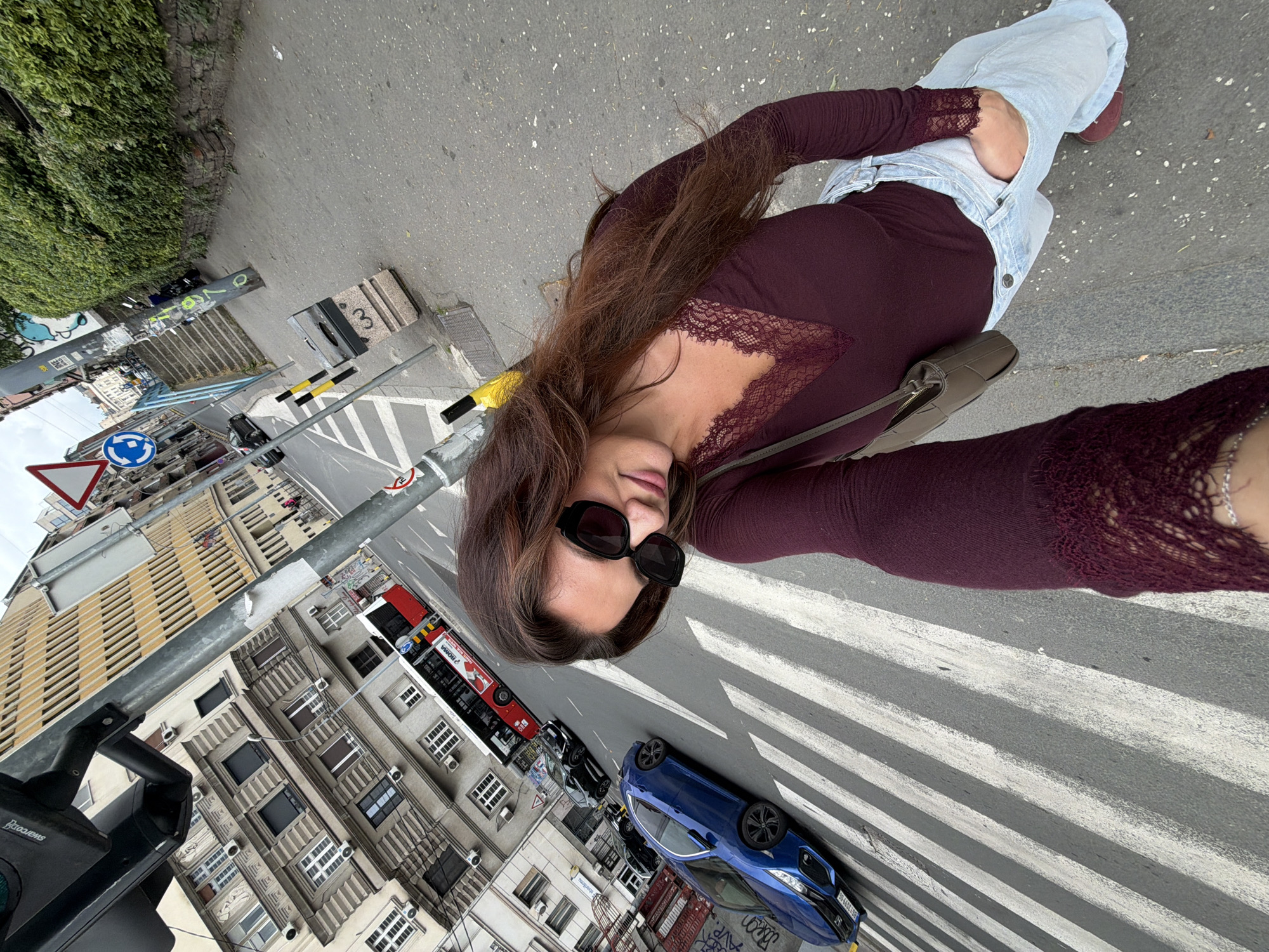 A woman is standing on a pedestrian crosswalk in Belgrade, Serbia, taking a selfie. The crosswalk is marked with white stripes. A blue car is parked nearby on the street. In the background are buildings, a bus, a street light, and road signs. The woman is wearing sunglasses, a burgundy long-sleeved shirt with lace trim, light blue jeans, and brown shoes. She also has a purse slung over her shoulder. The street signs and buildings suggest an urban environment.