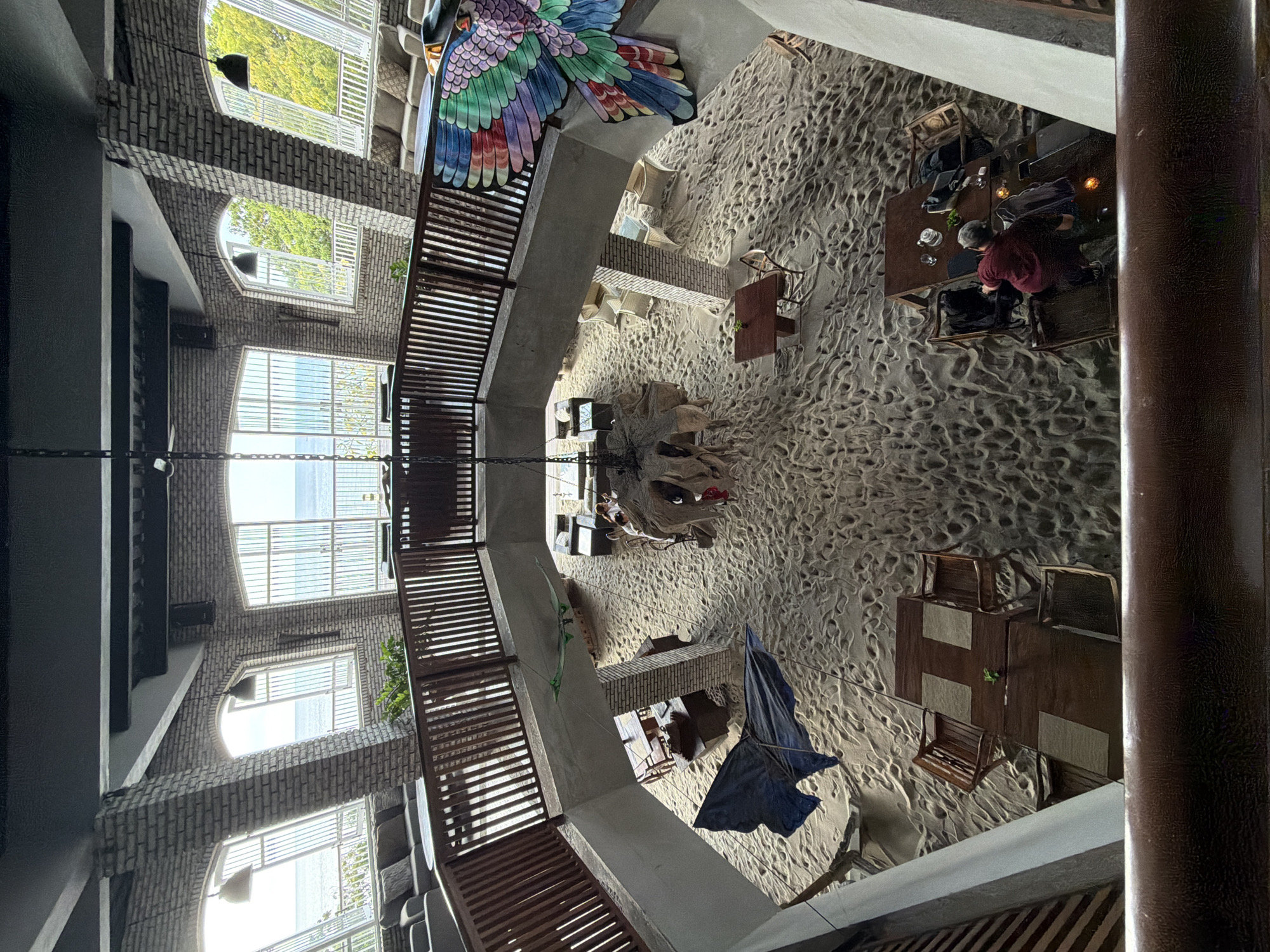 The image shows an indoor dining area with sand covering the floor, resembling a beach. The space has a mezzanine level with a wooden railing overlooking the main area. Large windows on the far wall offer a view of the ocean. Several tables and chairs are set up on the sand. In the foreground, a railing is visible. A large, colorful bird decoration is mounted on the wall to the right. On the mezzanine level, seating areas with sofas are arranged near the windows. In the main dining area, there are various wooden tables and chairs. A blue tarp is partially visible on the sand. Two people are seated at a table on the lower level, with one person in a maroon shirt leaning forward.