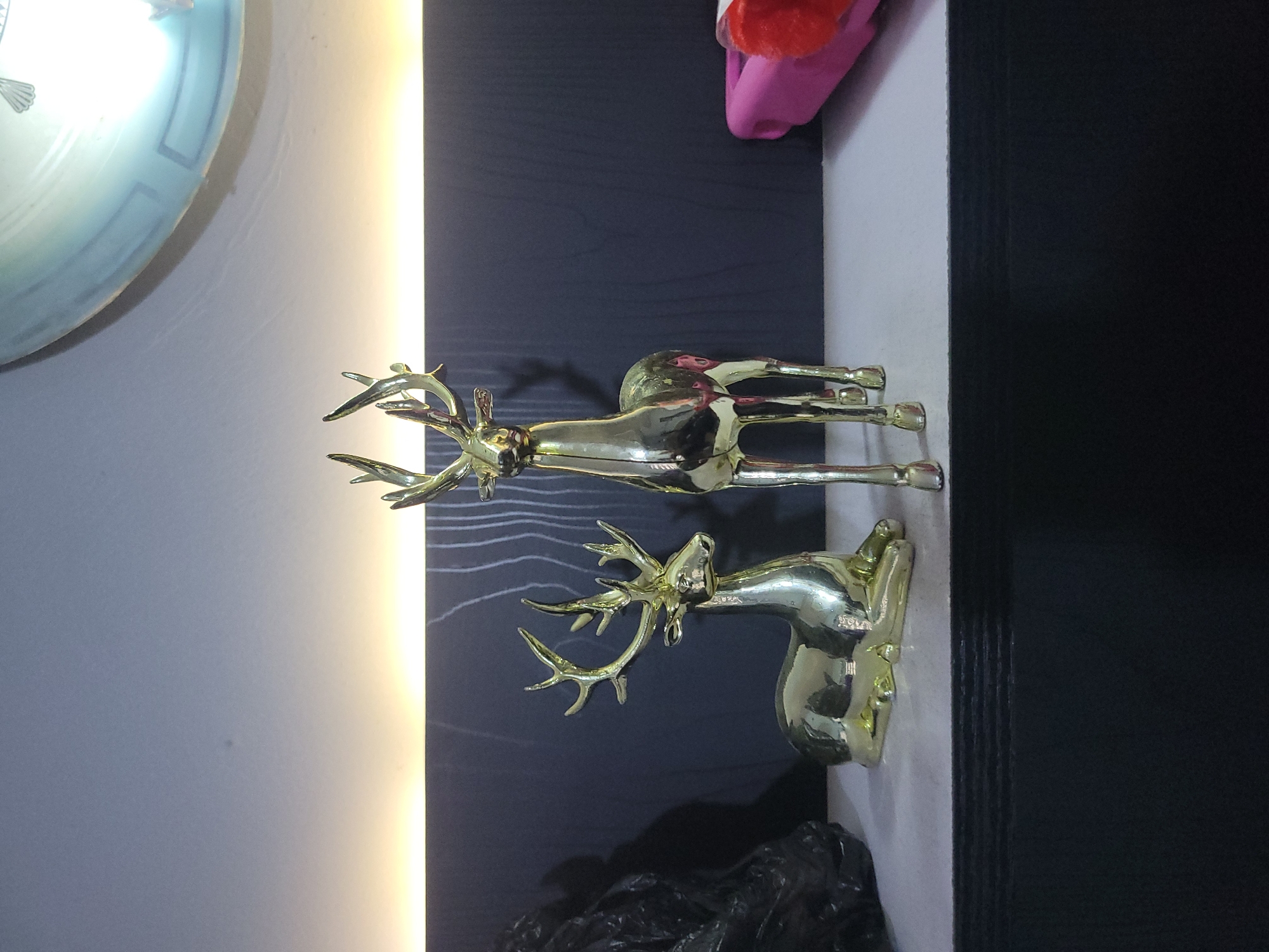Two golden deer figurines are displayed on a shelf. One deer is lying down, while the other is standing. The background features a dark, textured wall with a horizontal strip of warm light above it. A large, circular object, possibly a clock or a decorative plate, is partially visible in the upper right corner of the image. To the left of the deer, a dark, crinkled object, resembling a discarded plastic bag or a crumpled piece of fabric, is present. To the right of the standing deer, a bright pink object with a red fuzzy element is partially visible, possibly a piece of luggage or a container. The shelf itself is a light gray color, and it sits on top of a dark, layered surface. The lighting in the scene is dim, with a prominent warm light source creating highlights on the metallic deer figurines.