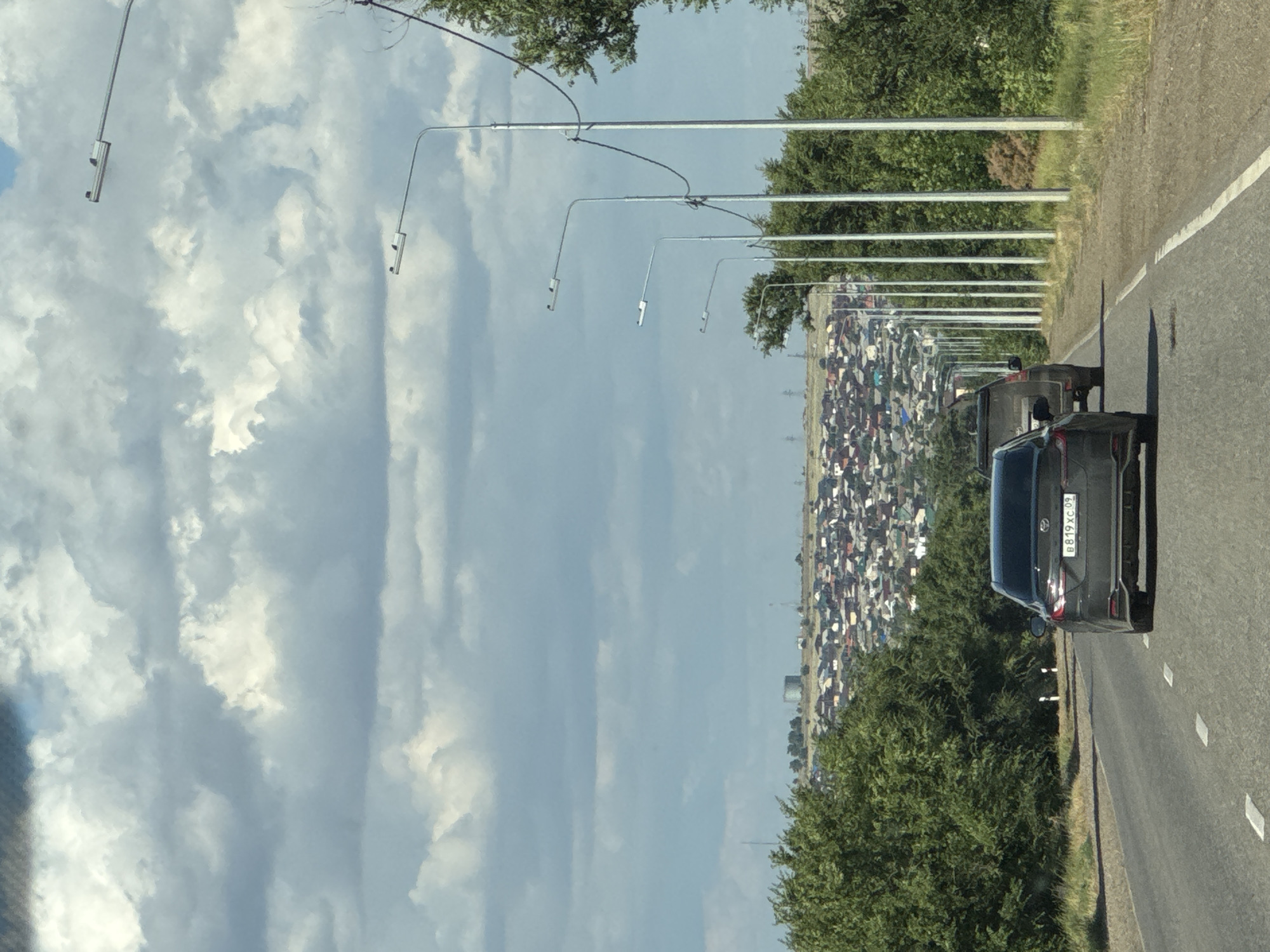 The media file shows a roadside view in Russia, likely during the daytime under a cloudy sky. The image is oriented vertically, with the road and a dense settlement of houses on the right side, and a large expanse of cloudy sky on the left.
The primary subjects are the road, a line of streetlights, a settlement of numerous houses with red and gray roofs, and a dark-colored car. The car, visible in the foreground on the right, has a license plate with the text "B819XC 09". The houses appear to be clustered together, forming a village or a suburban area. Green trees are present along the side of the road and also within the settlement.
The scene captures a snapshot of traffic on a road adjacent to a residential area. The activity taking place is implied to be driving or being a passenger in a vehicle. The lighting suggests it is daytime, possibly late afternoon or morning, given the presence of shadows and the overall brightness. The weather appears to be partly cloudy, with significant cloud cover in the sky.
Notable details include the repetitive pattern of streetlights along the road, the density of the housing settlement, and the license plate on the car. The image conveys a sense of everyday life in a Russian settlement with transportation infrastructure. There are no explicit emotional cues, but the scene depicts a common roadside view.