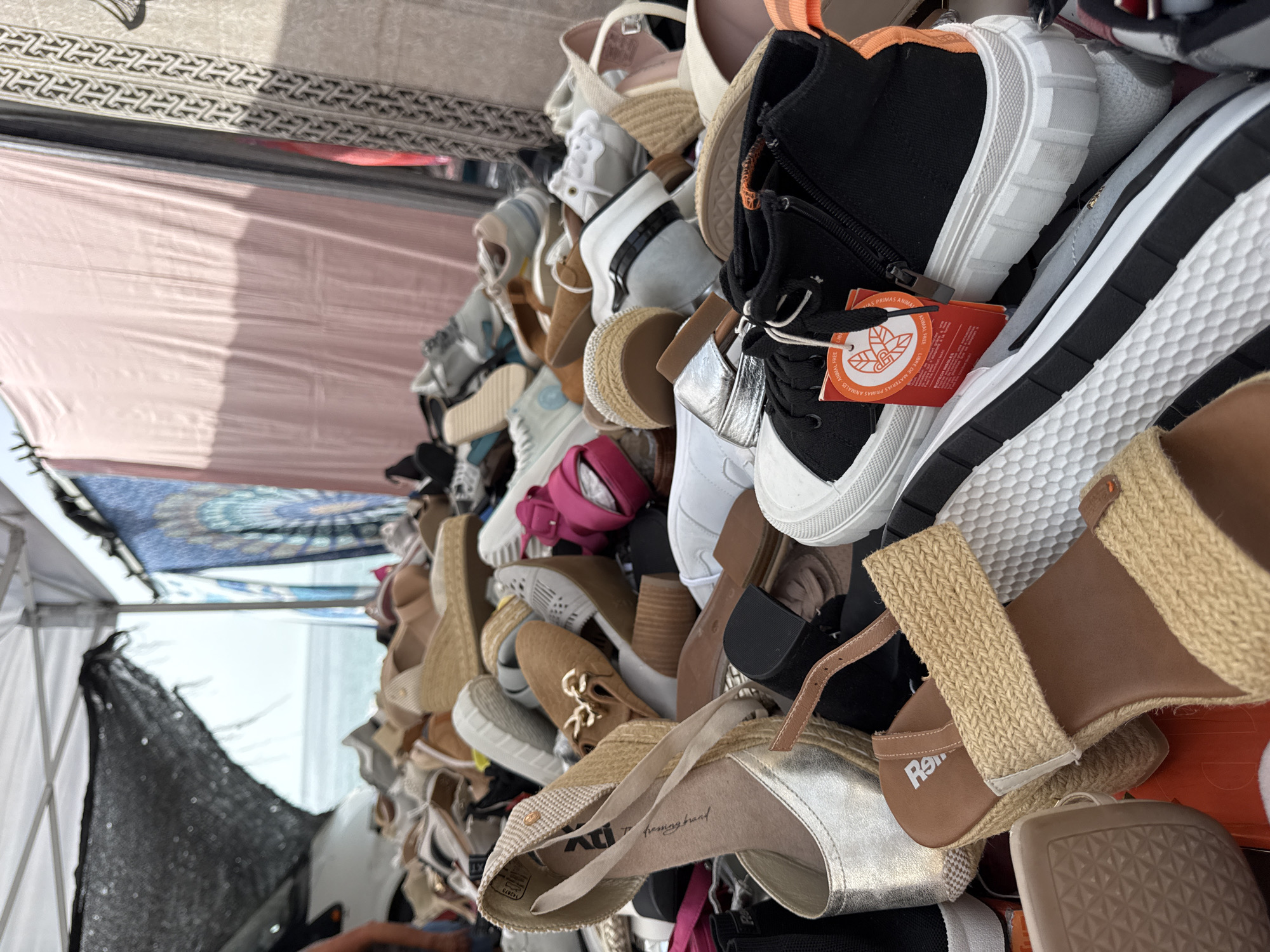 This image captures a close-up, low-angle view of a pile of footwear for sale at an outdoor market, possibly along the coast in Blanes, Spain, given the hint of the sea and sky in the background. The foreground is dominated by a variety of shoes, including espadrille wedges with woven raffia straps, a silver wedge sandal, black high-top sneakers with an orange accent and a red tag, and brown slip-on shoes with gold embellishments. Other shoes, including sneakers and sandals in various colors and styles, are visible in the midground and background, forming a dense collection.
The market appears to be set up under a white canopy, with draped fabric in pink and blue visible behind the shoes, suggesting a stall or tent. A glimpse of the sea and sky through a mesh screen in the upper left corner suggests the market is located near the coast. The overall impression is one of a bustling, open-air retail environment focused on footwear. There are no clearly discernible people in the frame, though the presence of a stall implies vendors. The lighting suggests it is daytime, likely with bright sunlight casting shadows.
