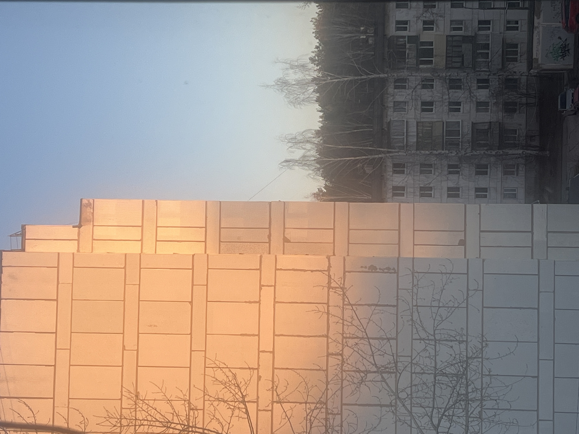 The image shows two buildings in the city of Chaikovsky, Russia. The building in the foreground is made of concrete blocks, with a pattern of vertical and horizontal lines. Part of the building is reflecting a sunset, casting an orange glow. Bare tree branches are visible in front of the building. Behind this is a derelict apartment building with many windows. The building seems abandoned, with faded paint and signs of disrepair. Bare trees are in between the two buildings. The sky is a pale blue.