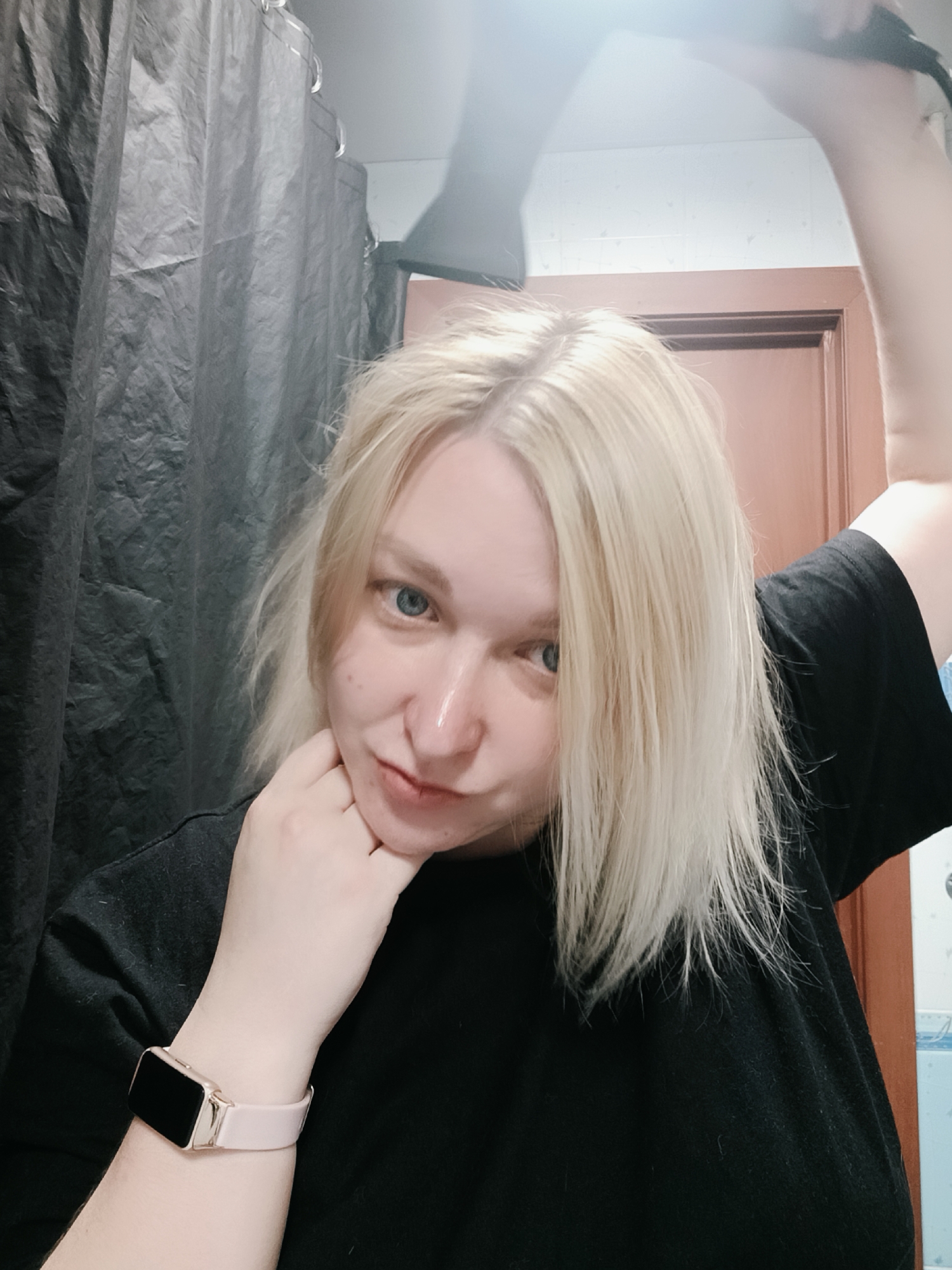 A woman with blonde hair and blue eyes is holding a black object above her head in a bathroom. She is wearing a black shirt and a pink smartwatch. Behind her is a black shower curtain and a brown door. The location is Chaikovsky, Russia.