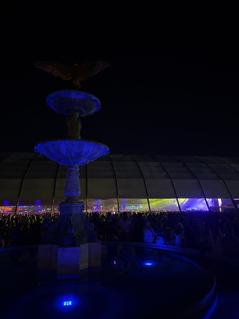 The image depicts a vibrant night scene at a large outdoor event, likely a music festival or concert, in Indio, United States. In the foreground, a prominent, ornate multi-tiered stone fountain serves as a central subject. It is illuminated by striking blue lights that highlight its details and the water within its basins. Perched on the top tier of the fountain is a sculpture of a large bird, possibly an eagle, with its wings outstretched.
Behind the fountain, a dense crowd of people fills the wide area, extending into the mid-ground. They are gathered under a massive, arched structure, resembling a large tent or stage canopy, which stretches across the background. Beyond the crowd and beneath this canopy, bright and colorful stage lights, primarily in yellows, greens, and blues, are visible, indicating a performance or stage area.
The time of day is clearly night, evidenced by the dark sky and the reliance on artificial illumination. The blue lighting of the fountain creates a cool, atmospheric effect, contrasting with the more vibrant and varied stage lights in the distance. The scene captures the scale and energy typical of a large gathering or festival. There is no visible text in the image.