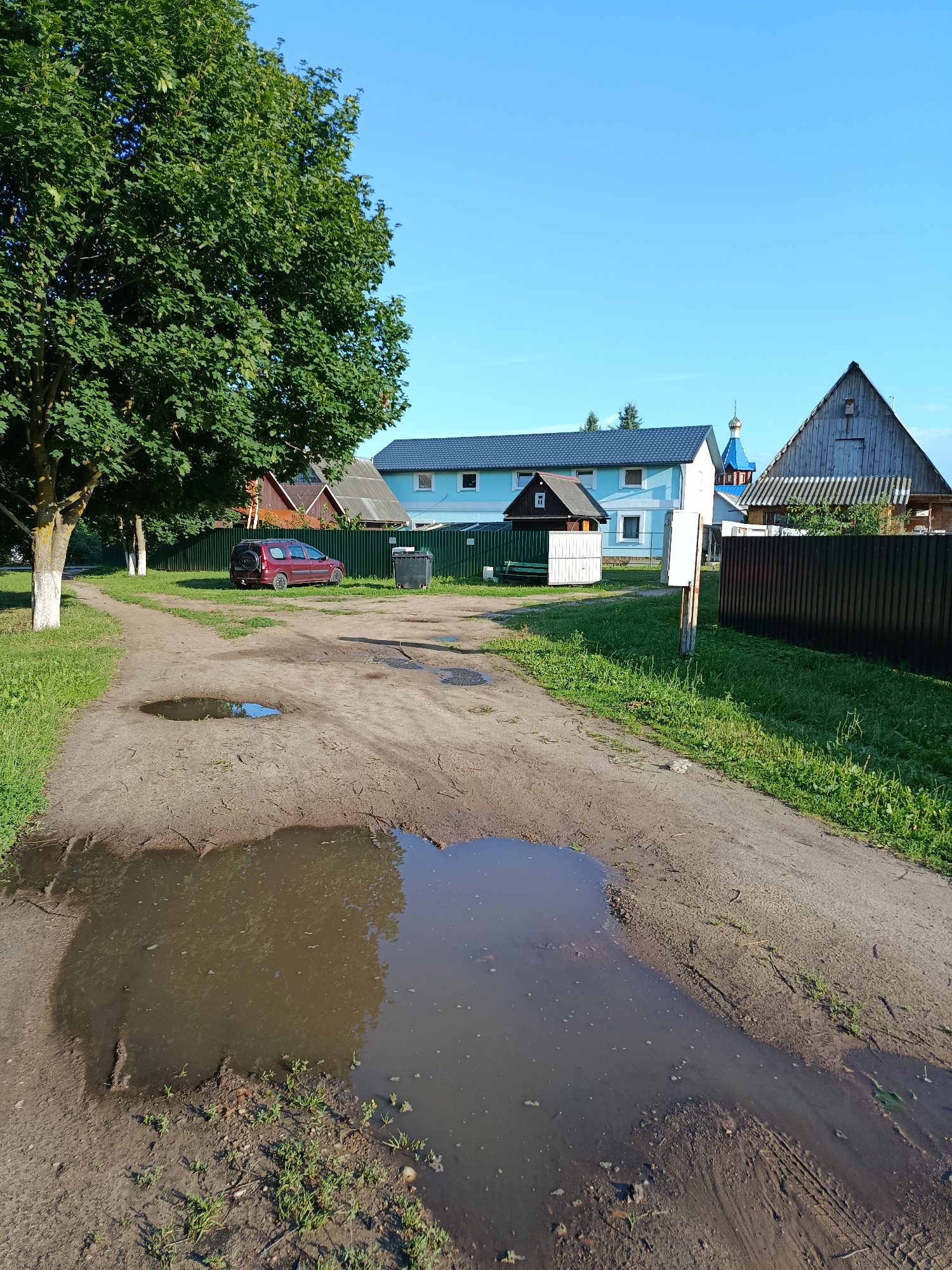 A dirt road with large puddles of water runs through a rural area under a clear blue sky. A large tree with green leaves stands on the left side of the road, and a red car is parked on the right. In the background, there are several houses, a fence, and what appears to be a church with a blue dome and cross. The scene is quiet and appears to be a typical day in a Belarusian village.