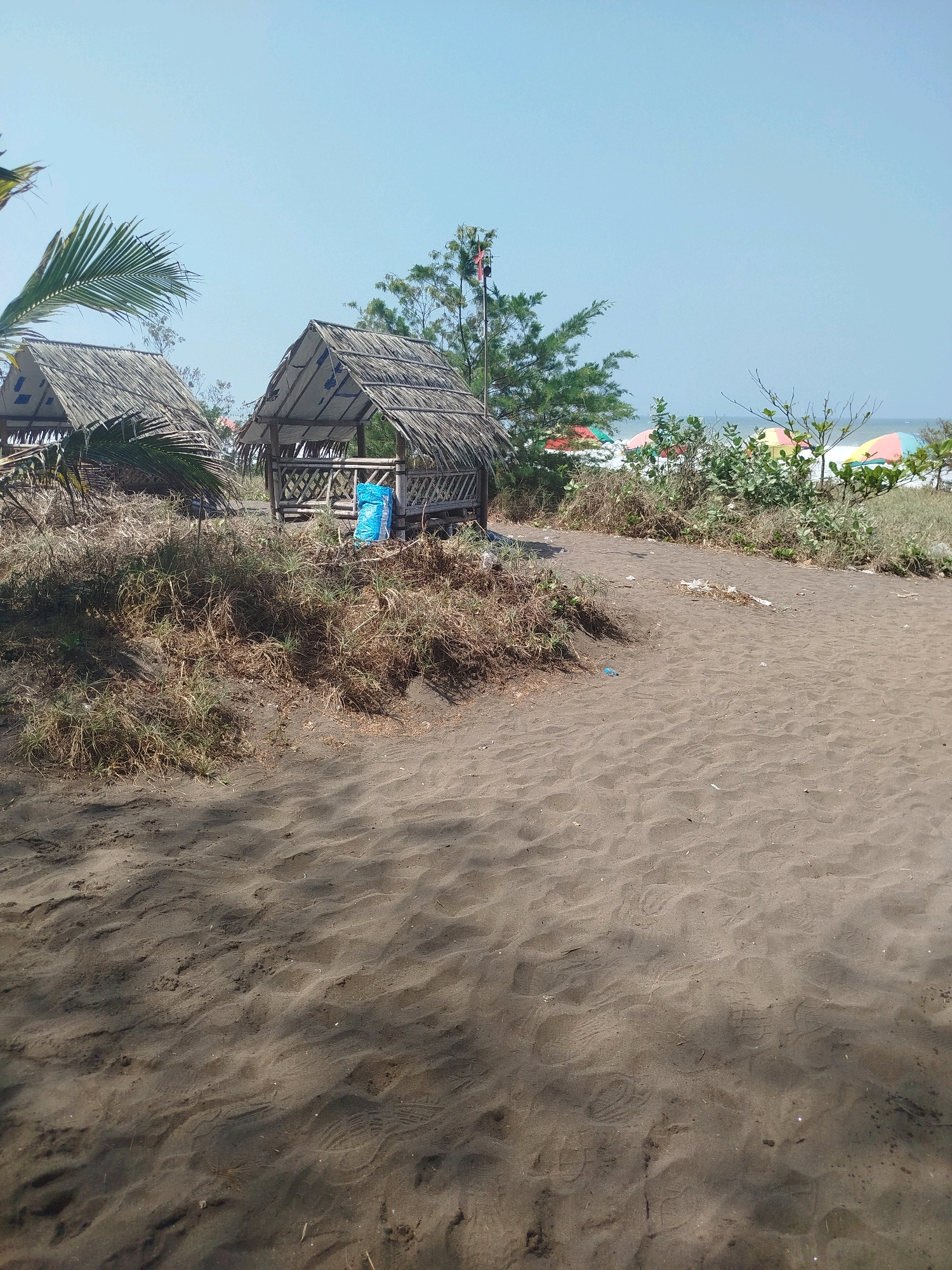 The image depicts a sandy beach scene with two small, rustic huts constructed with thatched roofs and bamboo walls. One hut is partially obscured by palm fronds. The ground in the foreground is covered in dark sand with visible footprints. Behind the huts, dry grasses and some greenery are present. In the background, lush vegetation and several colorful beach umbrellas can be seen bordering the ocean under a clear blue sky. A flag on a pole is visible behind the huts. The overall impression is that of a quiet, possibly somewhat undeveloped, beachfront area in Kenoyajaya, Indonesia.