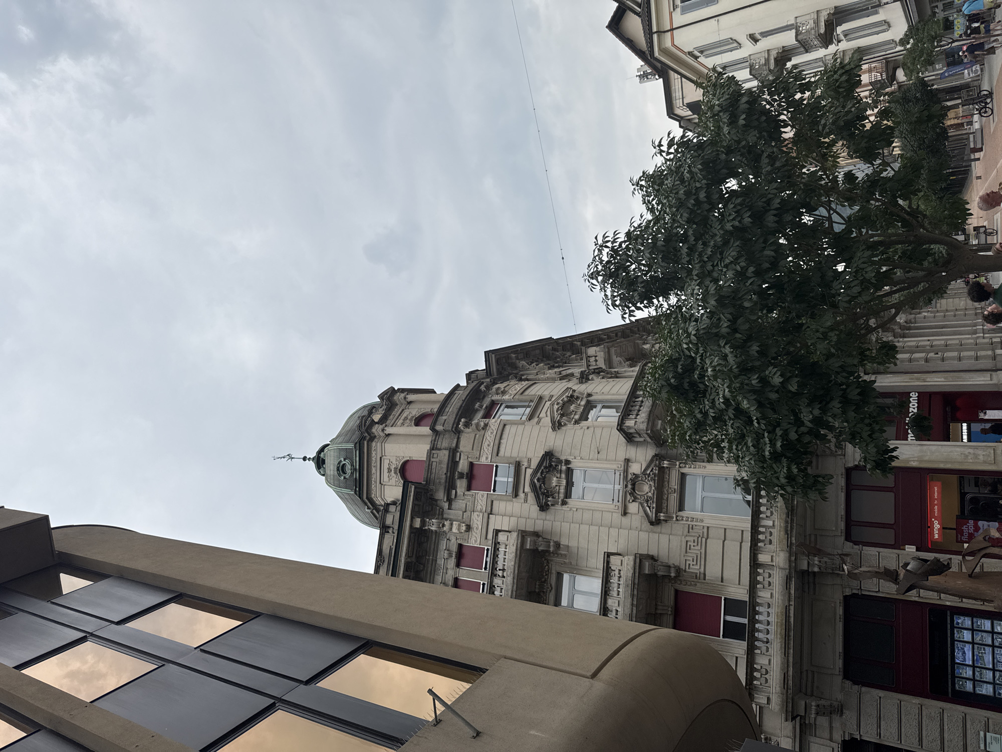 The image captures a view of the Via Carlo Maderno in Bellinzona, Switzerland, on an overcast day. On the left, a modern building with curved beige cladding and dark framed windows reflecting a warm sky is visible. To its right, a grand, older building with a green dome and intricate stone facade dominates the scene. This building features multiple floors with arched windows and red shutters, ornate architectural details, and balconies. In front of these buildings, a large, leafy green tree stands, its branches extending towards the sky. The street level reveals storefronts, including one with a red awning and signs for "wingo" and "mobile tv internet." Further down the street, other buildings with balconies and arched windows line the pavement, where a few people are walking and bicycles are parked. The sky is filled with grey clouds, suggesting diffused light.