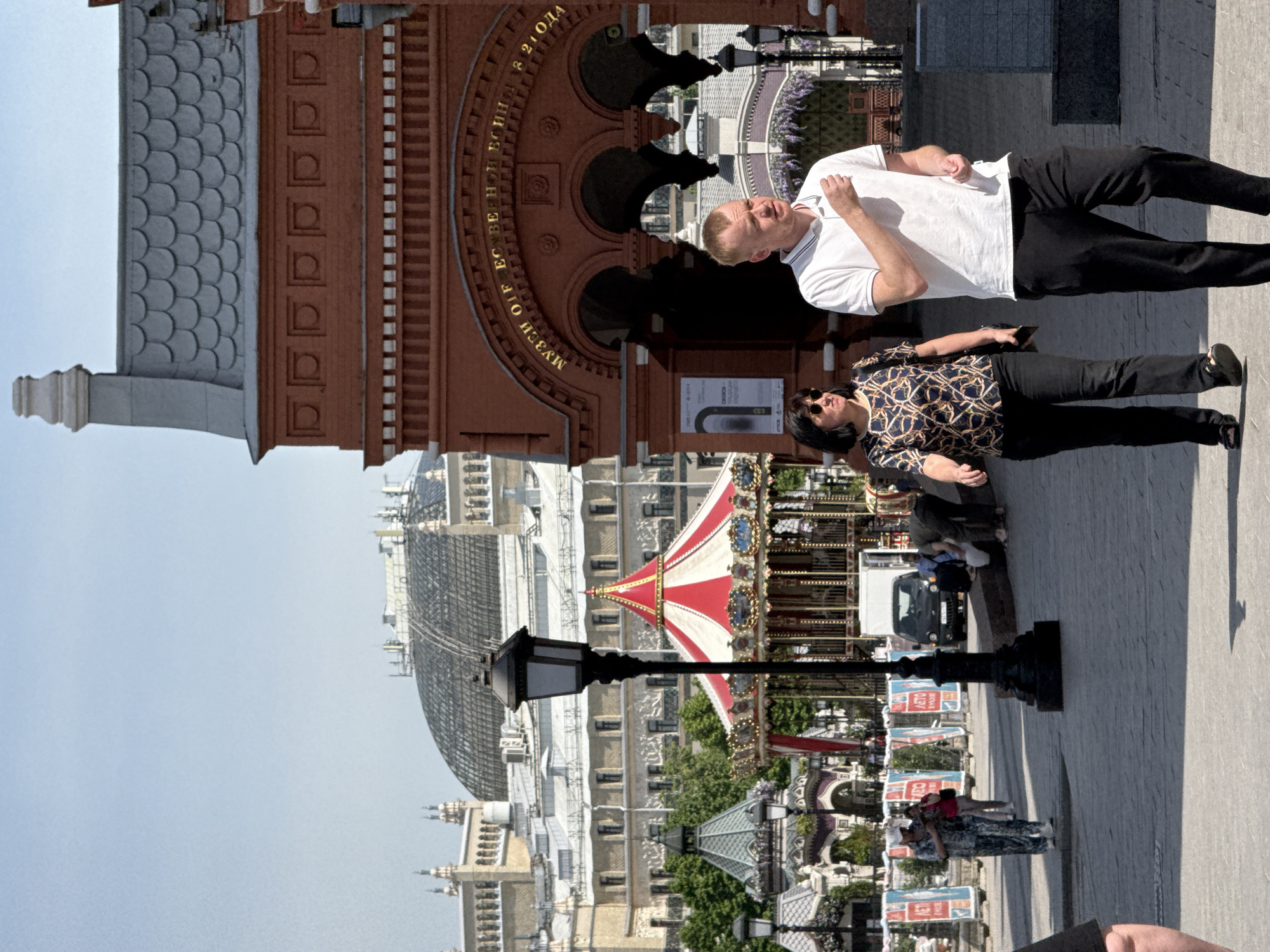 The image depicts a scene in Moscow, Russia, featuring the State Historical Museum and a carousel. The museum's facade, made of red brick with arched windows, and the museum name in gold lettering are visible in the upper left portion of the image. A carousel, striped red and white, stands near the center. A couple are in the upper right quadrant. The man wears a white shirt and dark pants; the woman wears a printed shirt and dark pants. The image is shot from a low angle, emphasizing the height of the buildings and the carousel. A lamppost stands in the foreground. The sky is light blue. Pedestrians are further down the street.