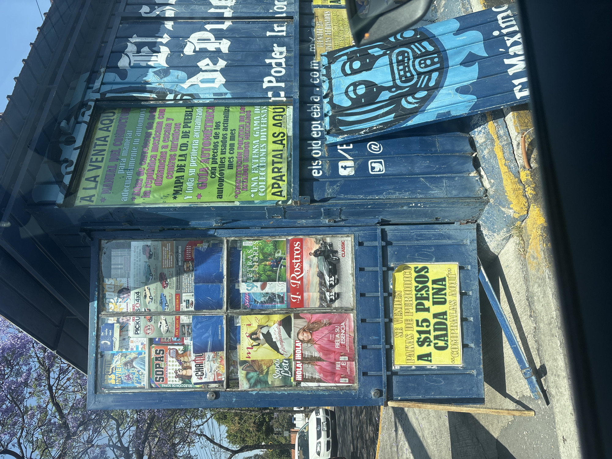 A street-side newsstand, likely in Puebla, Mexico, is photographed at an angle. The newsstand is constructed from a blue, metal container, painted with white graffiti-style writing, possibly political slogans. The structure features a display window showcasing magazines and newspapers. A yellow sign advertises "pakas de periodico" (packs of newspapers) for 15 pesos each. The side of the newsstand lists "A LA VENTA AQUI" (For Sale Here) with various manuals, maps, and guides, including a Puebla city map. Social media icons are also visible on the container. A partial view of street pavement and parked cars is seen below. Purple flowering trees are in the background.