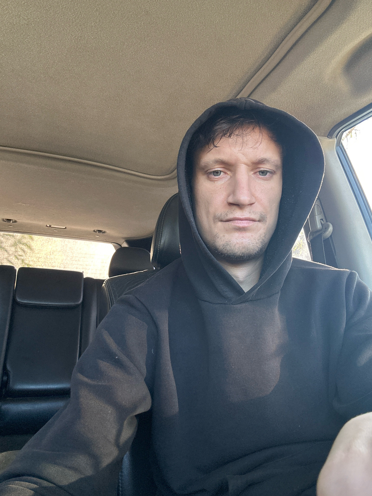 A man is pictured from the chest up, inside a vehicle. He is wearing a dark hooded sweatshirt, and the hood is up, partially obscuring his face. The man has light-colored eyes, brown hair, and stubble. The interior of the car appears to be made of dark leather and fabric. Sunlight streams in from the windows, suggesting it is daytime. The background shows the back seats of the car and the rear window. There is no visible indication of the specific location within the city of Makiv, Ukraine.