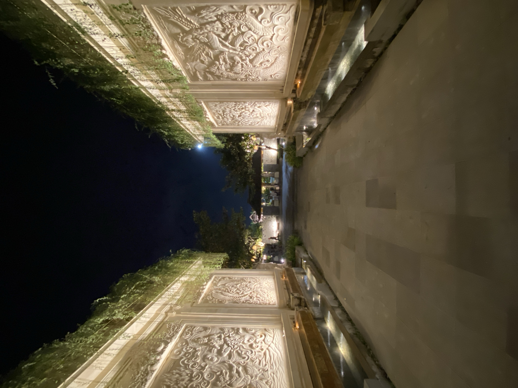 The image depicts a walkway at night in Kuta Selatan, Indonesia, likely within a resort or cultural complex. The walkway is flanked by two ornate, illuminated walls with intricate carvings and lush greenery cascading from above. The walls are lit from below, highlighting the detailed bas-relief sculptures, which appear to depict mythical creatures or traditional motifs. Small water features are integrated into the base of the walls, with gentle cascades of water adding to the ambiance. The walkway itself is paved with large, rectangular stones and is empty of people. In the distance, beyond the immediate walkway, there are trees and a structure with a traditional roof, also illuminated. The sky is dark, with a faint hint of moonlight visible. The overall impression is one of a serene and elegantly designed outdoor space at night.