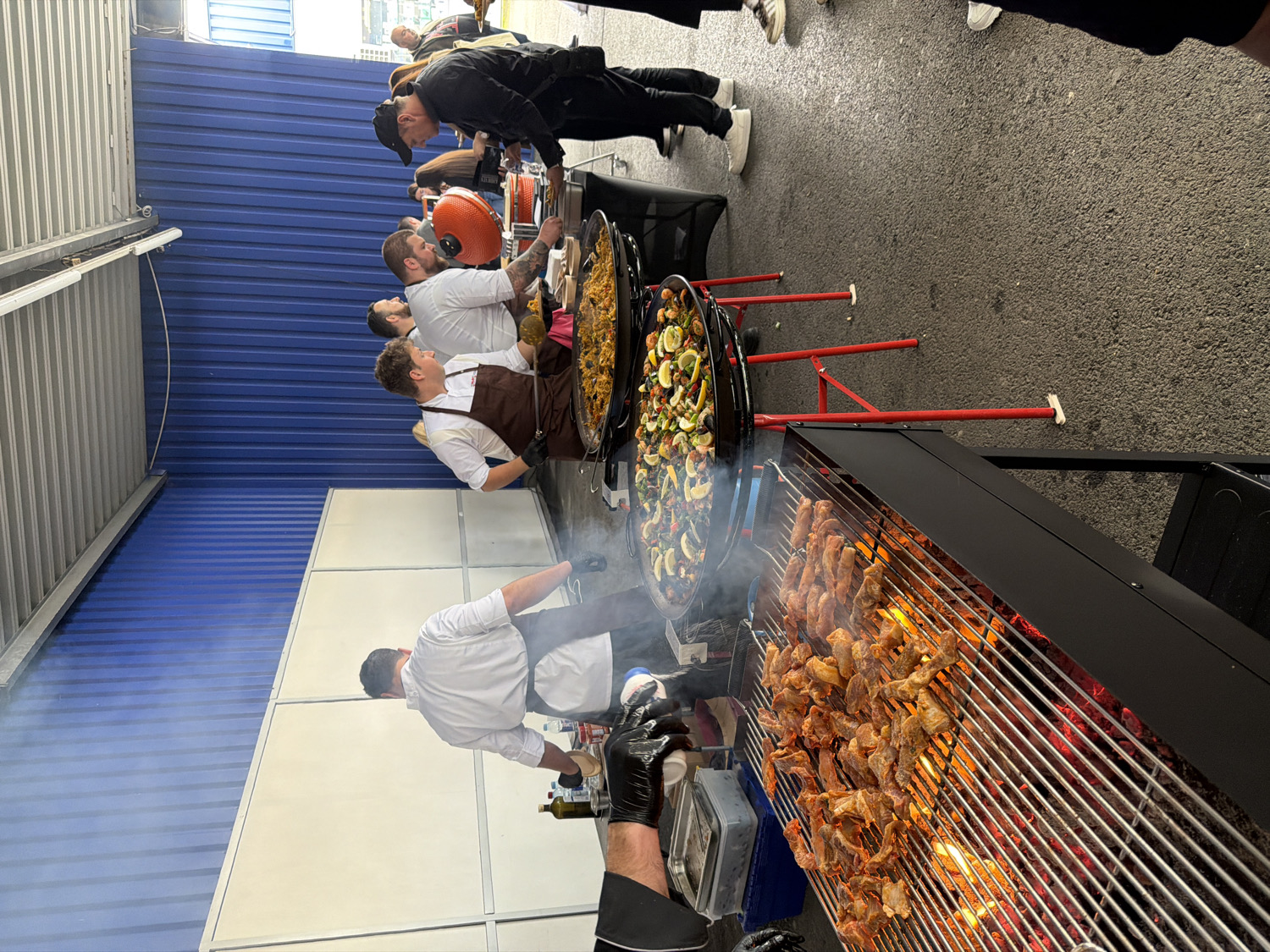 This media file captures an outdoor or semi-outdoor culinary event, showcasing several chefs preparing food on a large scale. The scene features multiple cooking stations set up against a backdrop of a blue corrugated metal wall and white paneling, with an asphalt ground.
In the foreground, a chef wearing a white coat is actively tending to a large grill, where numerous skewers of meat are cooking over open flames, producing noticeable smoke. His gloved hand is visible, suggesting he is adjusting or seasoning the food. Adjacent to the grill, further into the image, are two large paella pans resting on red metal stands. These pans are filled with food, including rice, seafood, vegetables, and lemon slices.
Several men, also dressed in chef attire (white coats and aprons), are gathered around the paella pans. One chef is actively stirring the contents of a pan with a large utensil, while another stands nearby, holding a spoon and observing the cooking process. A man in a black cap and jacket appears to be overseeing or supervising the operation near the paella pans. Other individuals are present in the background, observing the cooking activities.
The event seems to be a food preparation demonstration or catering service, possibly at a festival, market, or outdoor gathering, given the portable cooking equipment and the informal setting. The lighting suggests it is daytime. There is no clearly legible text visible in the image.