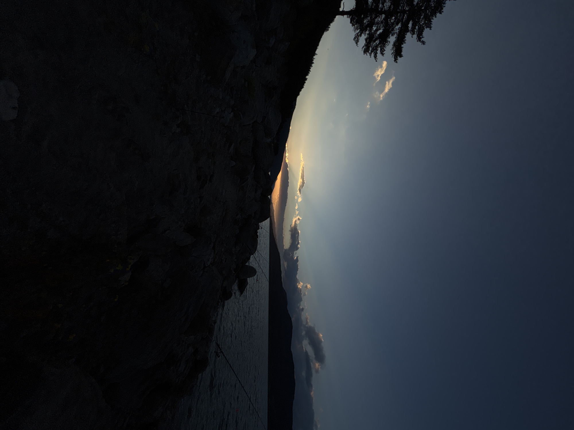 The image depicts a twilight scene at a rocky lakeside. A silhouetted evergreen tree stands on a steep, dark slope to the left, overlooking a body of water. The sky is a deep blue, transitioning to lighter shades near the horizon where the sun is setting behind a dark mountain range. Wispy clouds catch the last rays of sunlight, appearing as streaks of orange and white. The foreground is dominated by dark, rugged rocks and sparse vegetation, indicating a natural, undeveloped shoreline. Two fishing rods are visible planted in the rocks near the water's edge, suggesting recreational fishing. The overall atmosphere is serene and tranquil, characteristic of a peaceful evening by the water.